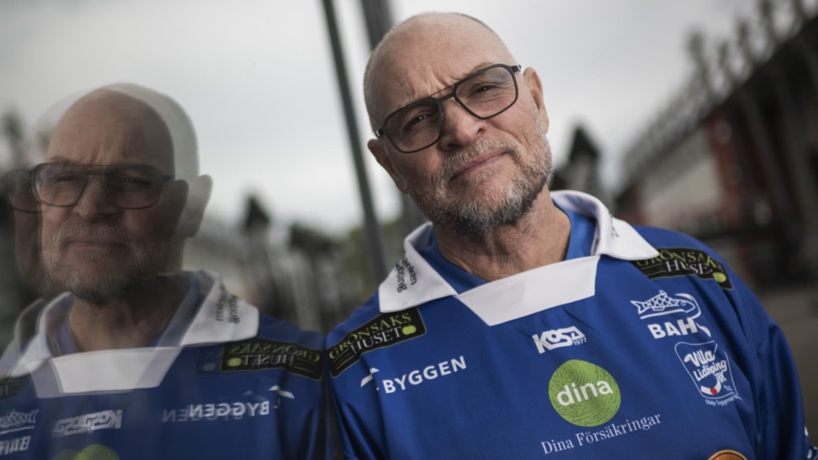 Två personer klädda i blåa sporttröjor med sponsorloggor står utomhus vid en brygga. Bakgrunden visar en molnig himmel och suddiga detaljer av en byggnad.