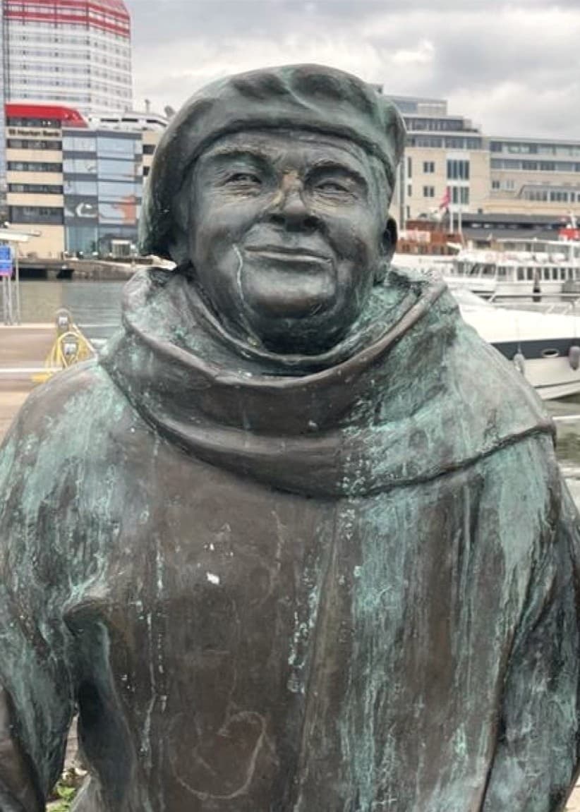 Staty av Evert Taube i brons vid vattnet med bakgrund av byggnader, båtar och molnig himmel. Statyn visar Taube i en tänkande pose med en fot vilande på en träbalk.