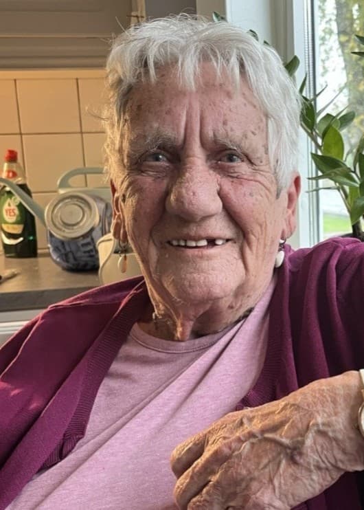 En äldre kvinna sitter i ett kök med en rosa tröja och vinröd kofta. Hennes hand med en vit klocka vilar på fönsterblecket.