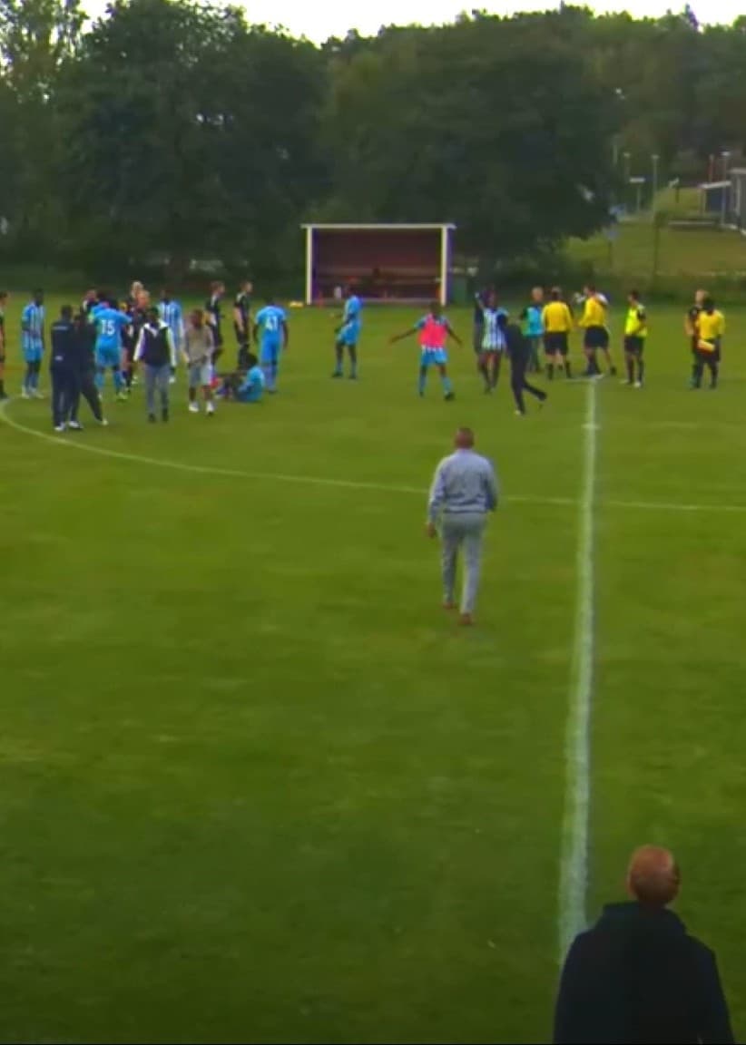 Fotbollsplan med spelare och domare samlade efter en skandalmatch, Anton Carlsson i svart jacka med klubbemblem står vid sidlinjen.