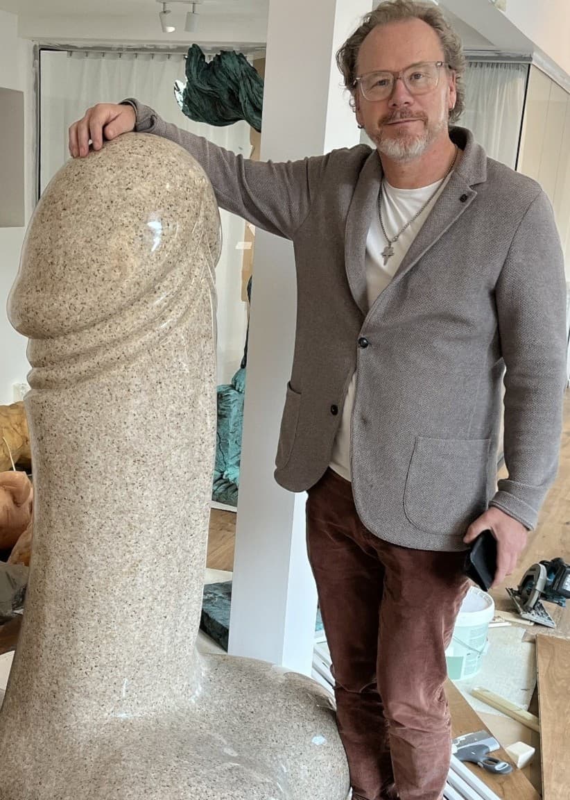 Marcus Skinnar står bredvid en stor granitskulptur i form av en penis, som han köpte i Kina för över 20 år sedan. Rummet är fyllt med andra konstverk och skulpturer.