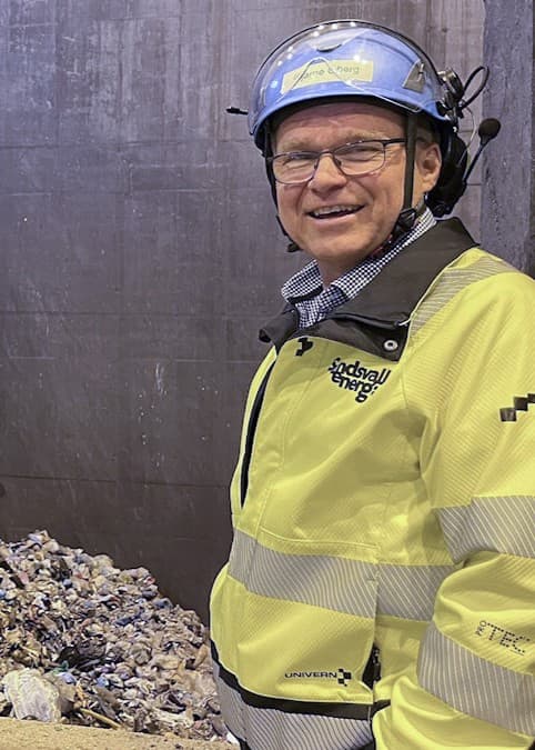 Till vänster syns en man i gul varseljacka och hjälm vid en stor hög av metallskrot. Till höger står en kvinna på en grusplan vid ett bostadshus med två stora svarta hundar.