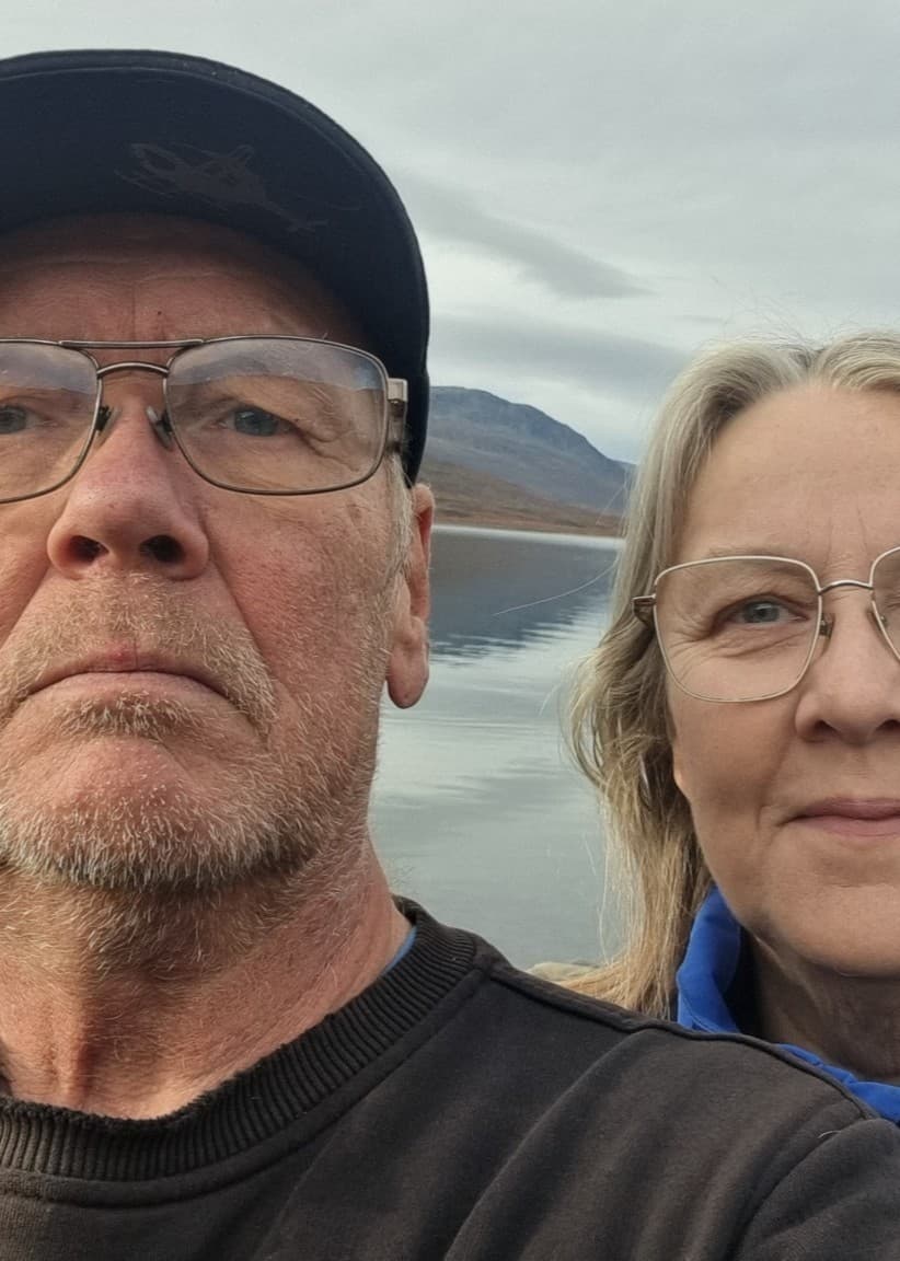 Två personer står vid en sjö med berg i bakgrunden. Den ena personen bär keps och den andra har långt hår. Landskapet domineras av gråa toner och en lugn vattenyta.