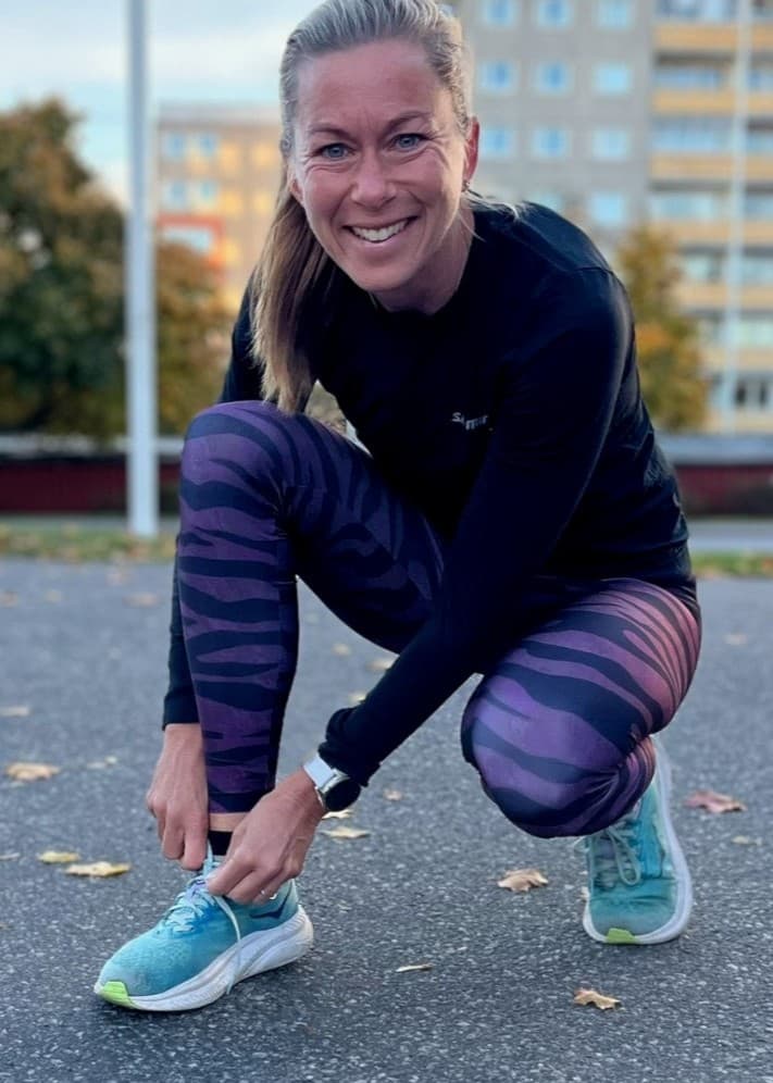 Frida Södermark, löpare och träningsinspiratör från Norrköping, ses knyta sina löparskor på en asfalterad yta. I bakgrunden syns höghus, höstfärgade träd och parkeringsplatser.