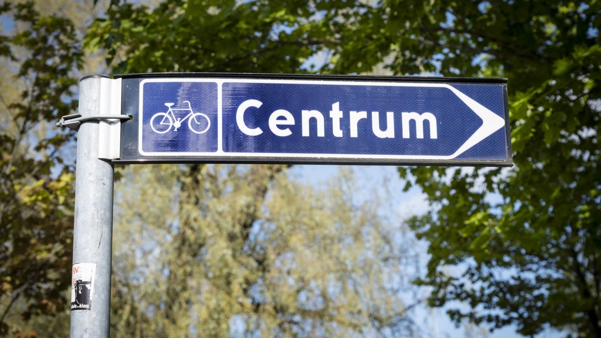 En blå skylt med texten 'Centrum' och en cykelsymbol visar riktning mot stadens centrum, omgiven av gröna träd och en klar himmel.