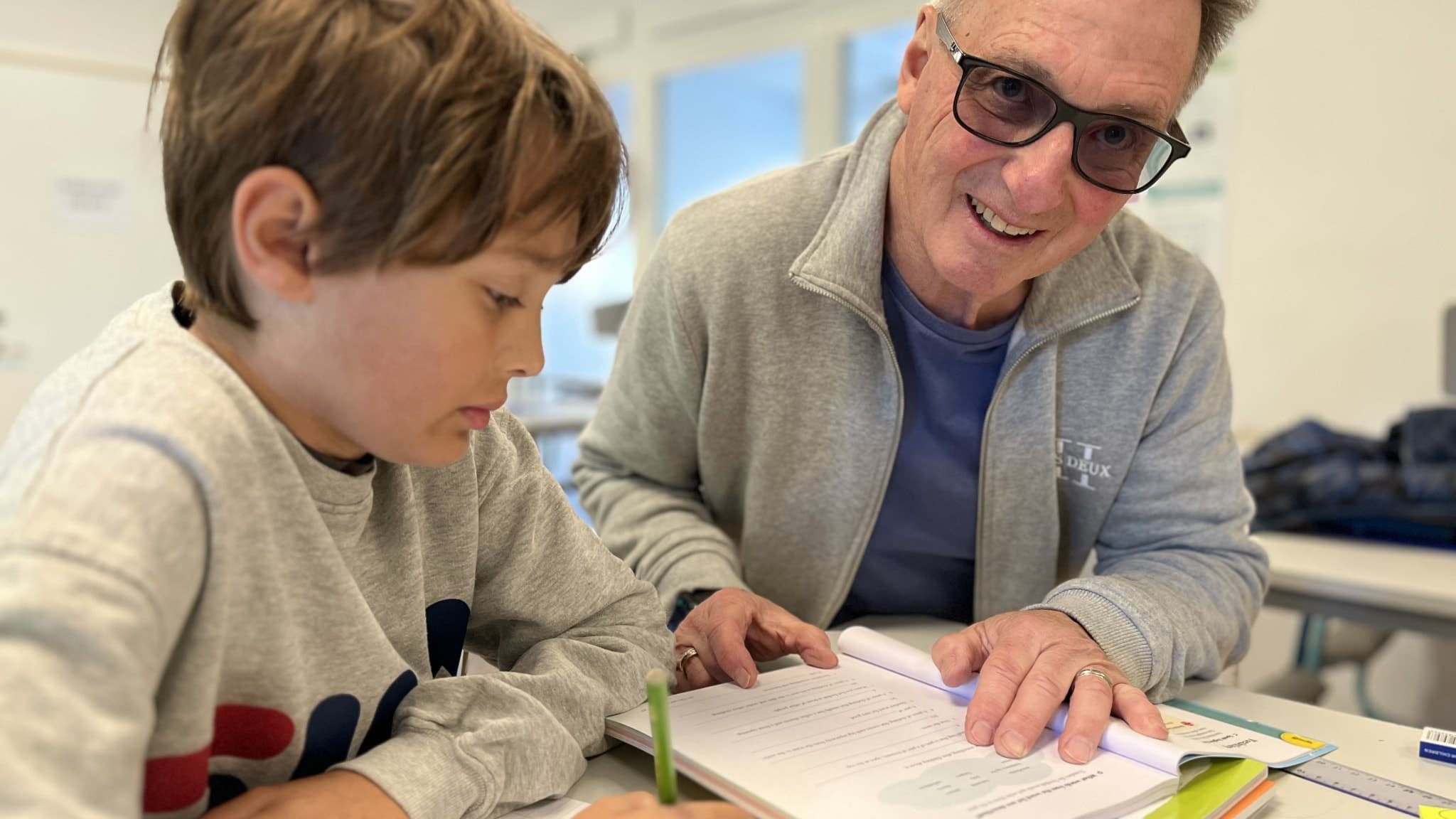 Peter, skolmorfar i Ekerö kommun, sitter vid ett bord och hjälper eleven Julian med skoluppgifter i ett klassrum.