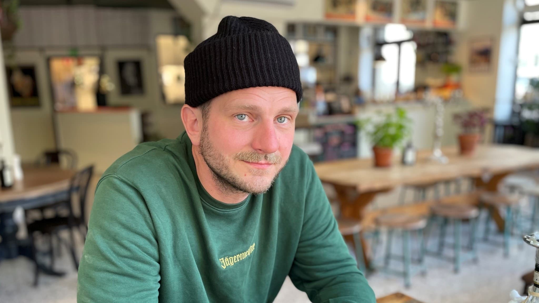En man sitter ensam i en restaurang. Mannen har en svart sotarmössa på huvudet och han bär en grön långärmad tröja. 