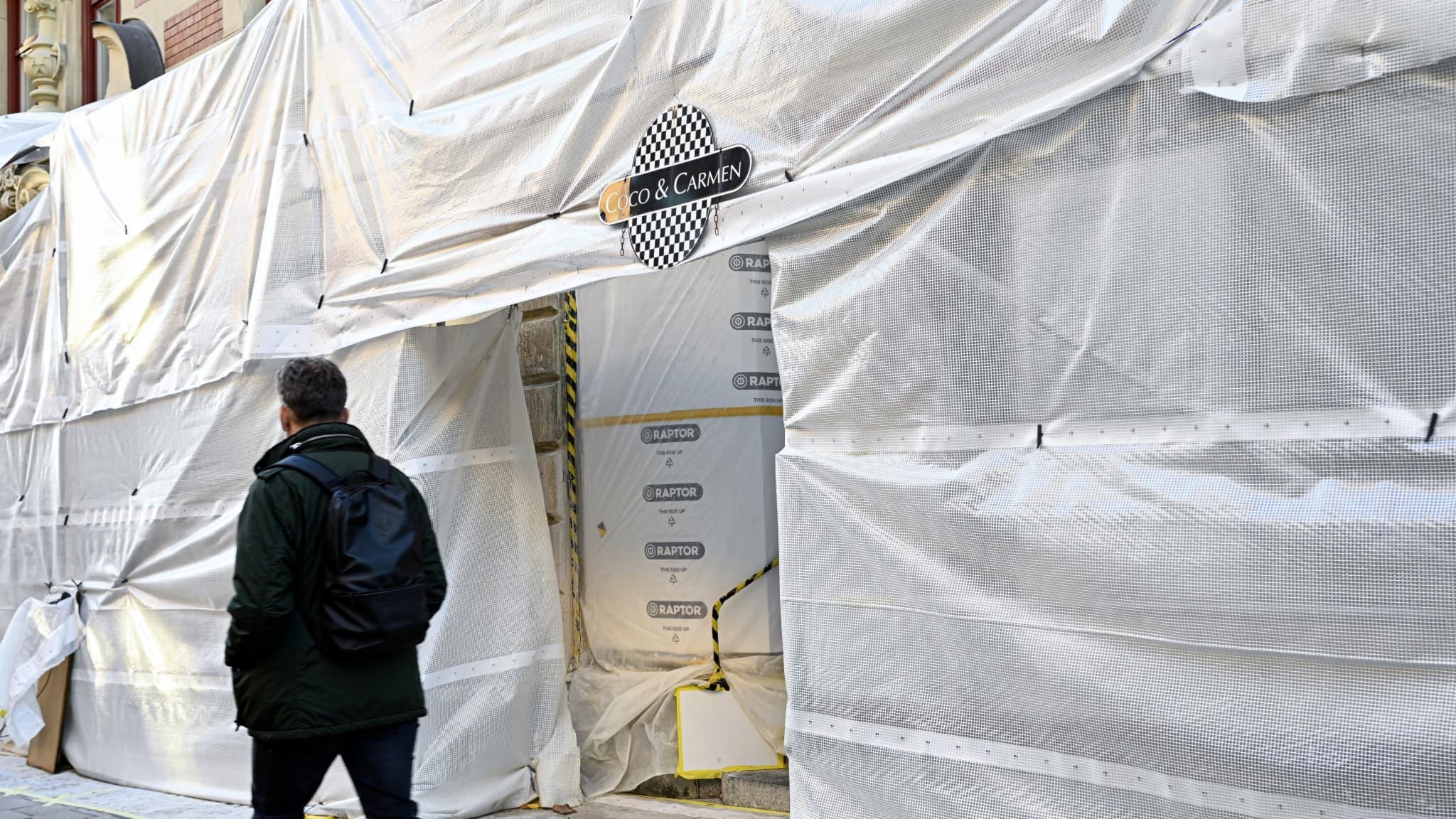 En person går förbi restaurangen Coco & Carmen på Östermalm i Stockholm, vars fasad är täckt av byggplast under renovering. Restaurangen kopplas till ett misstänkt listeriautbrott.