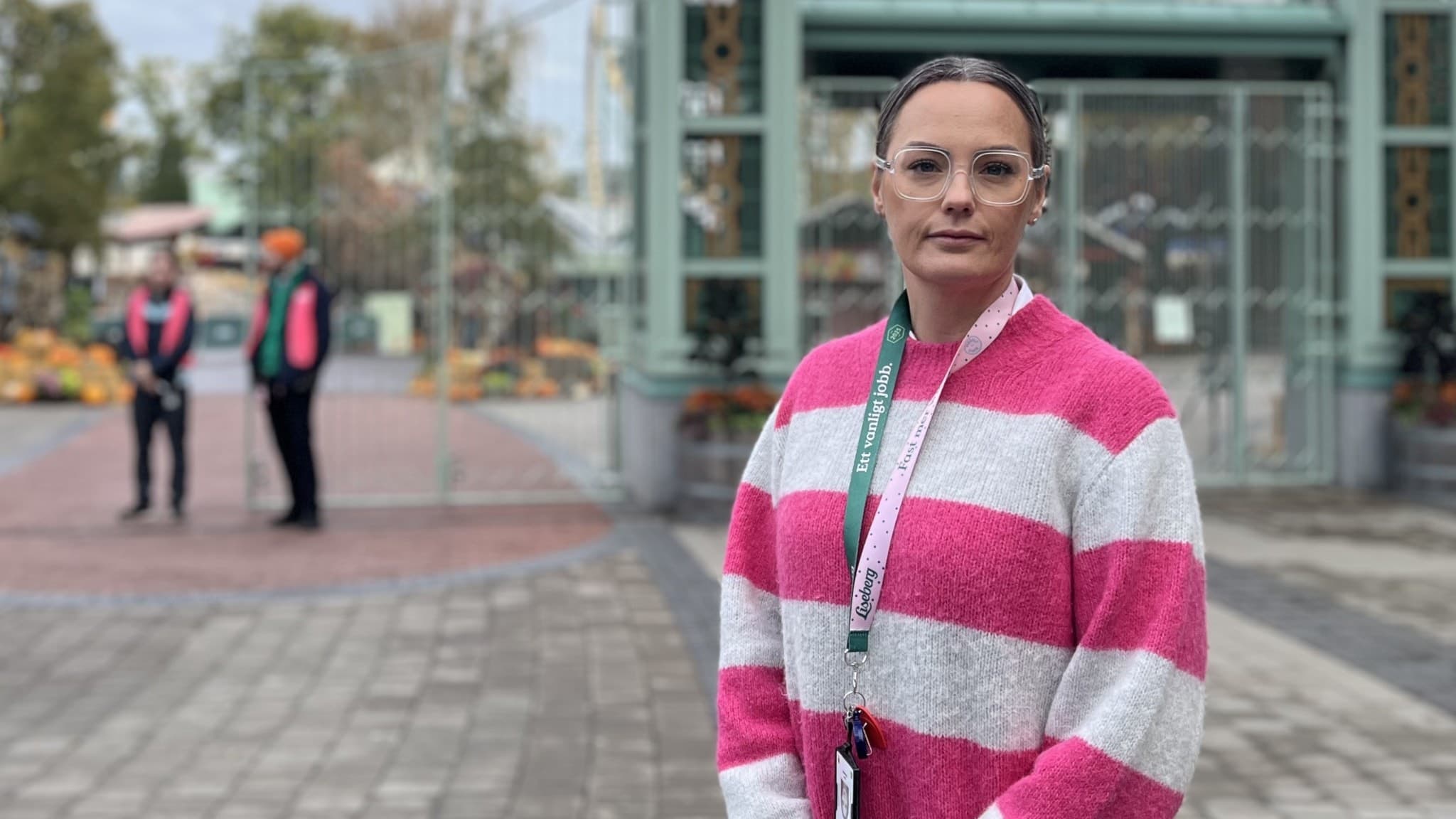 Clara Rosendahl står utomhus framför en glasentré, klädd i en randig rosa och vit stickad tröja och ett ID-band runt halsen. I bakgrunden ses två personer i uniform vid en entré.