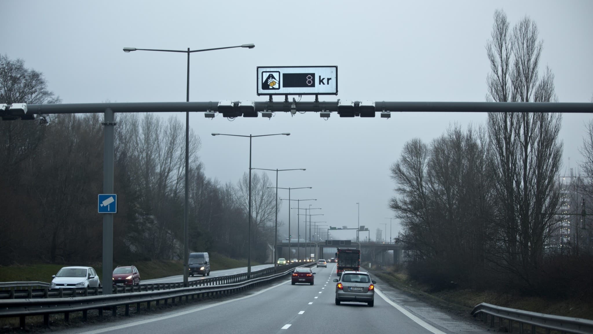 Motorväg i Göteborg med trängselskattsskylt som visar en avgift på 8 kronor. Flera bilar kör i båda riktningar under en gråmulen himmel, omgiven av träd och vägskyltar.