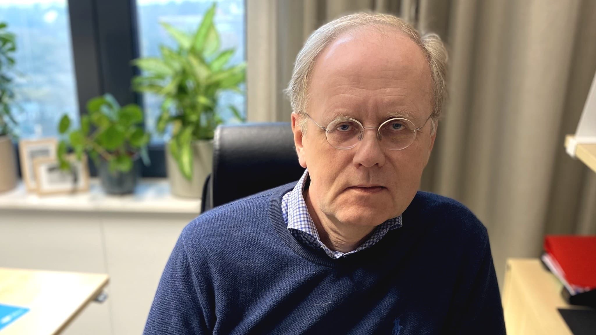 Smittskyddsläkaren Bengt Wittesjö sitter vid ett skrivbord i ett kontor med växter och fönster i bakgrunden, klädd i en mörkblå tröja över en skjorta.
