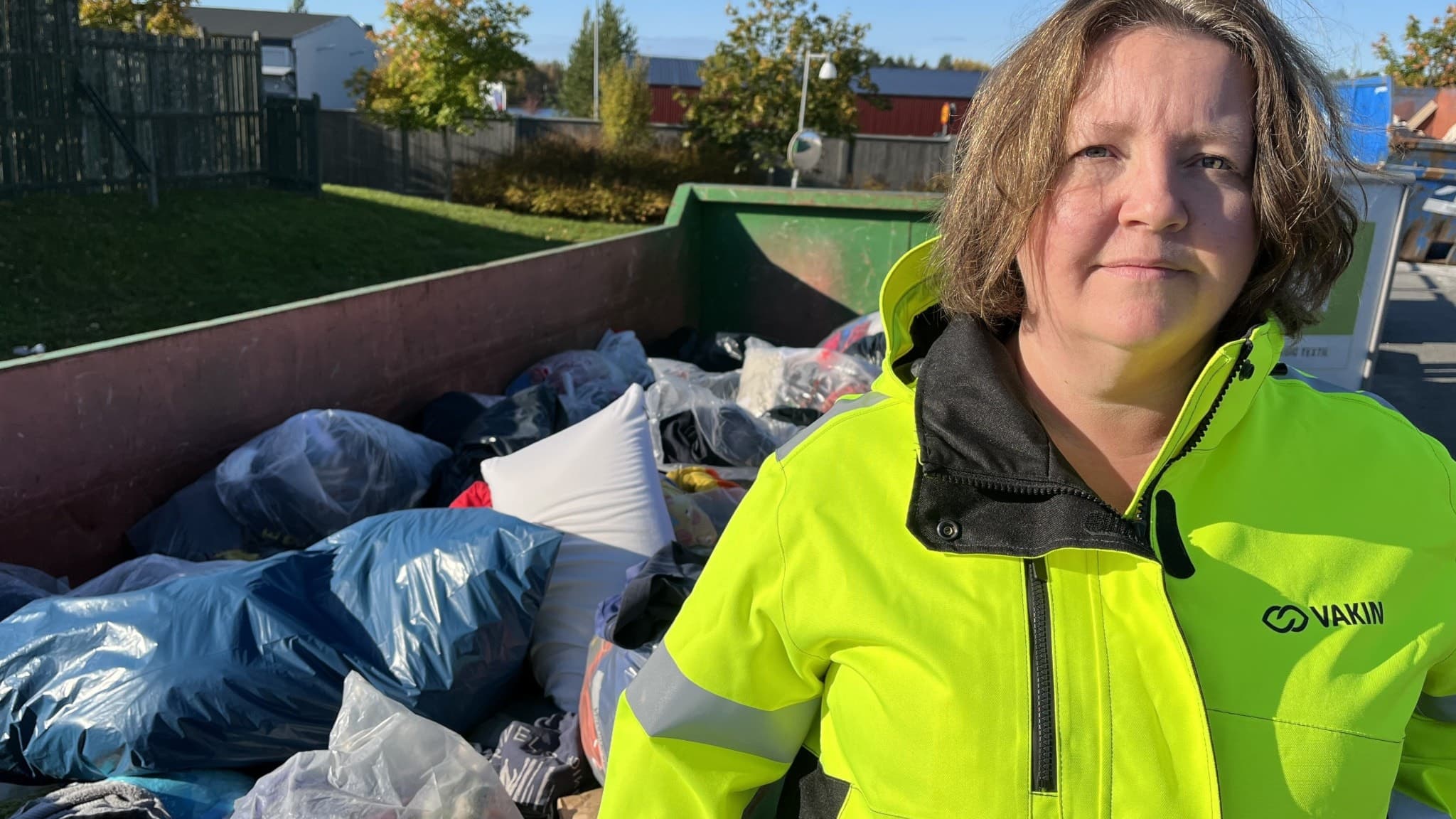 En person i gul arbetsjacka står framför en container fylld med textilier och andra föremål, utomhus i ett bostadsområde med träd och hus i bakgrunden.
