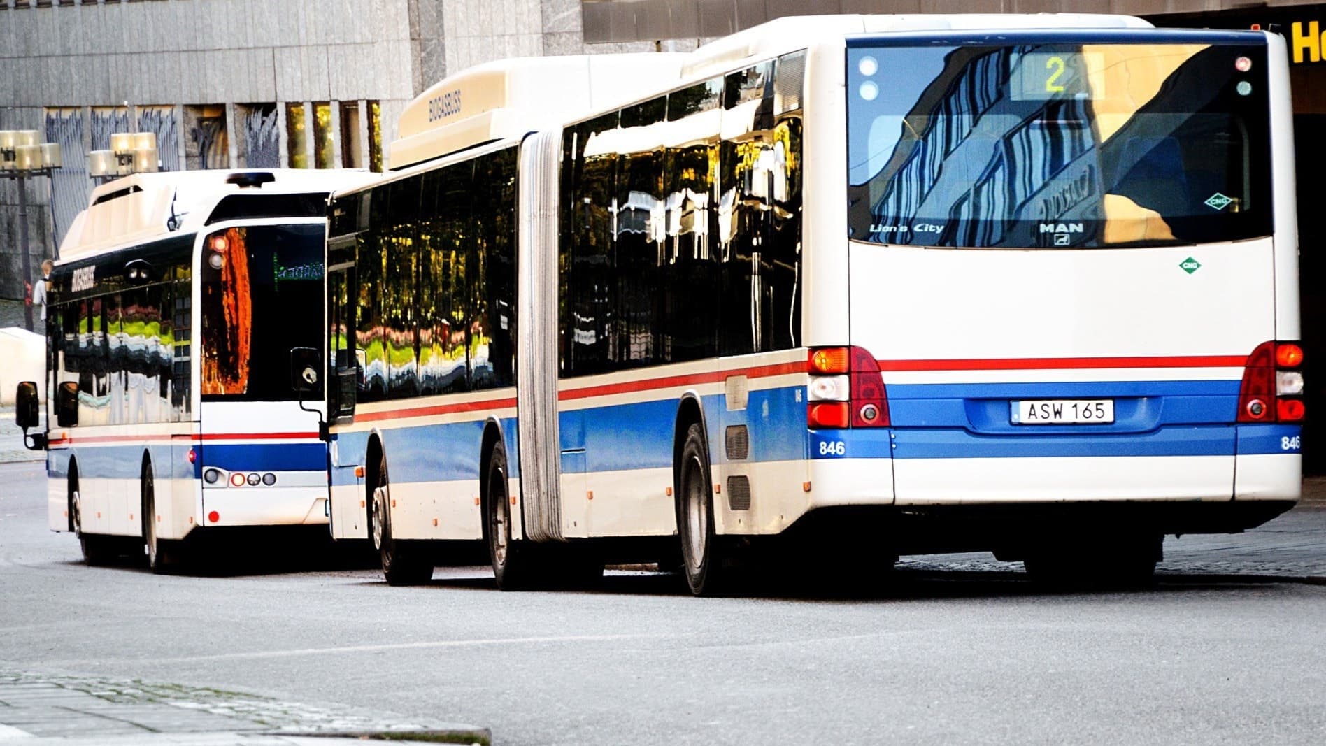 Två stadsbussar från Västmanlands Lokaltrafik står vid en hållplats i centrala Västerås. Bussarna är vita med blå och röda linjer.