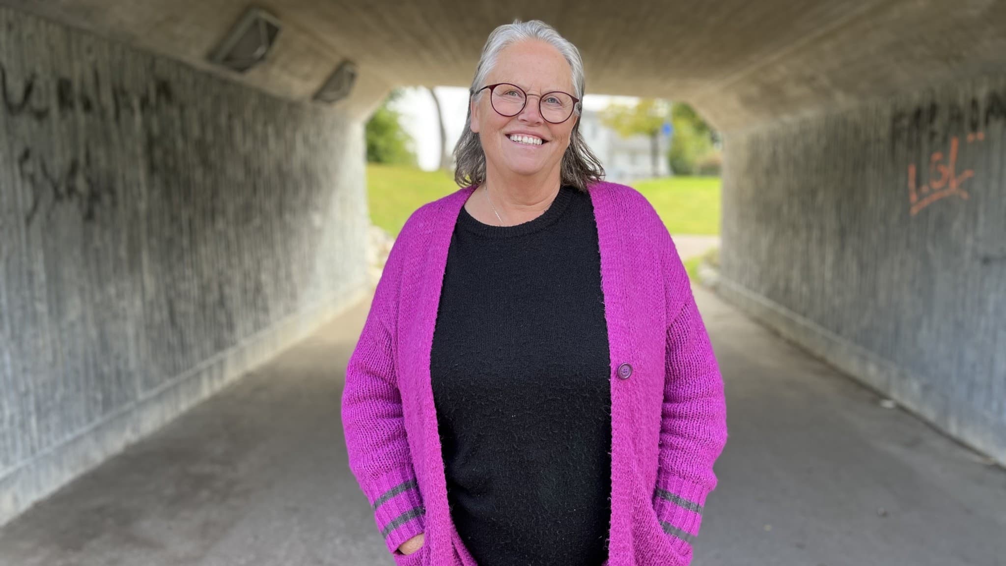 Vänsterpartisten Ann-Christine Andersson står i en gångtunnel med graffitimålade väggar, iförd en lila kofta och svart topp.