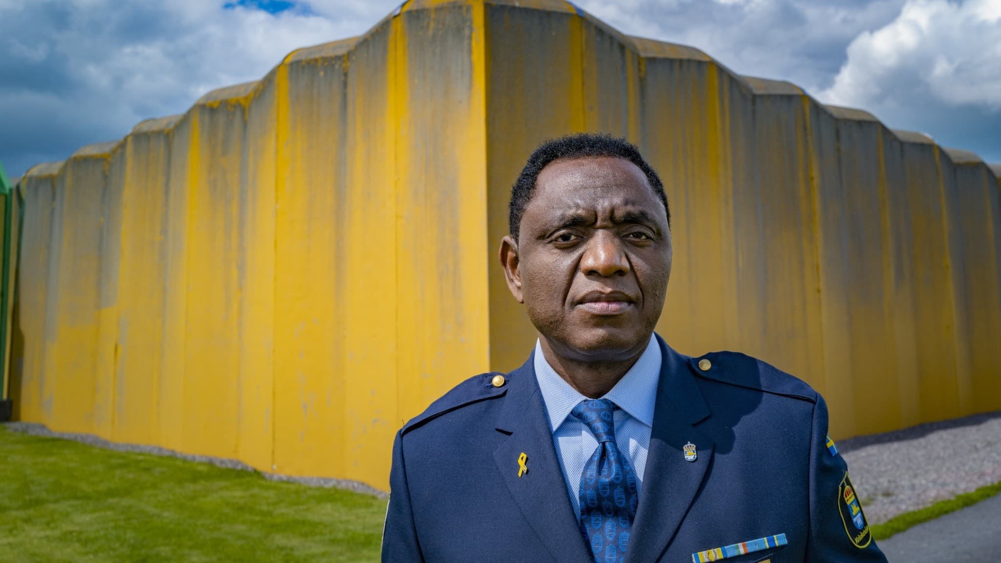 Jacques Mwepu, chef på Kumlaanstalten, står i uniform framför en gul byggnad med en molnig himmel i bakgrunden. Bilden fokuserar på säkerhet och fängelsemiljö.