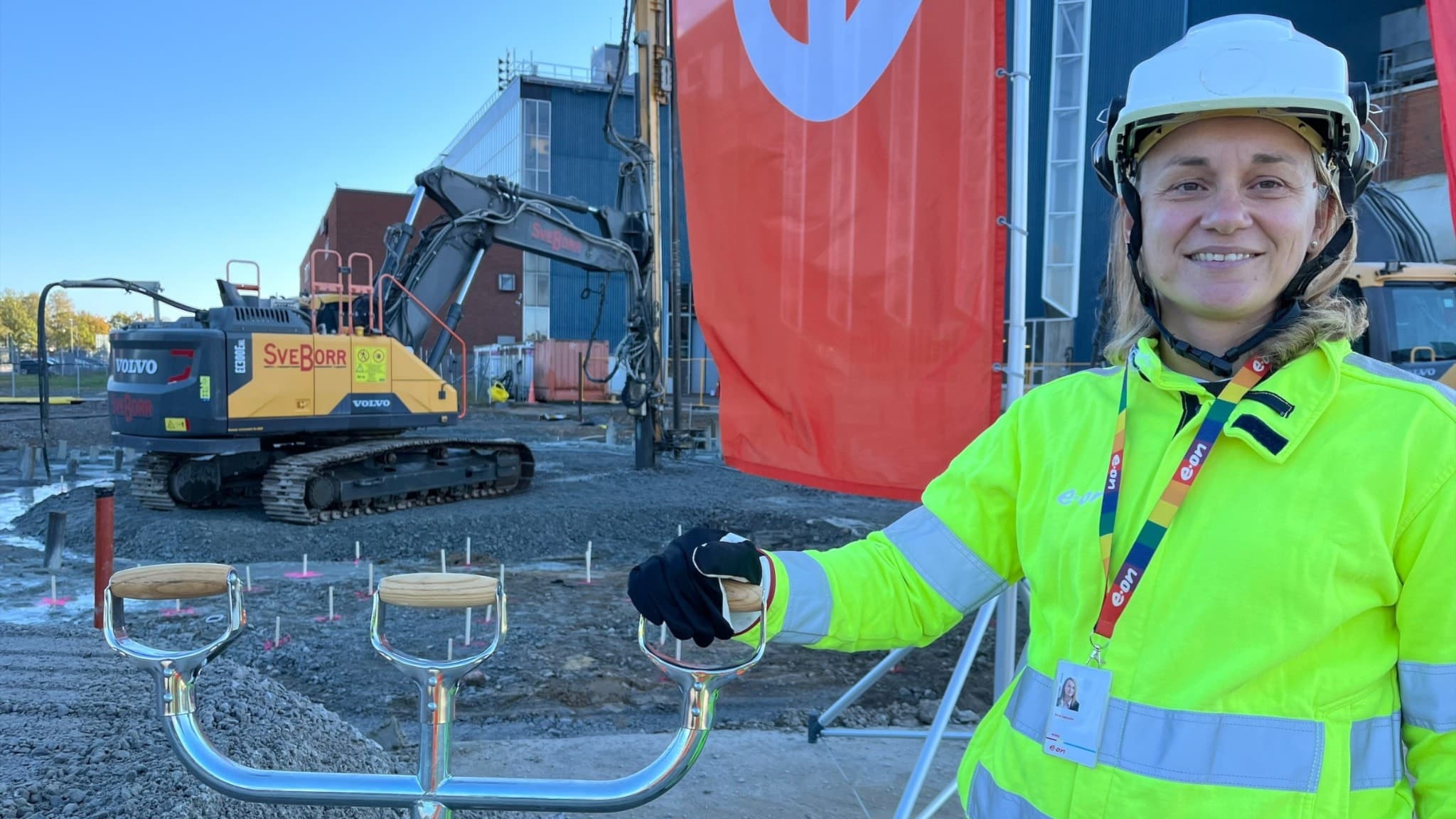 Azra Sapcanin står framför en byggarbetsplats med en grävmaskin och röda företagsflaggor i bakgrunden, iklädd gul skyddsjacka, hjälm och reflexer.