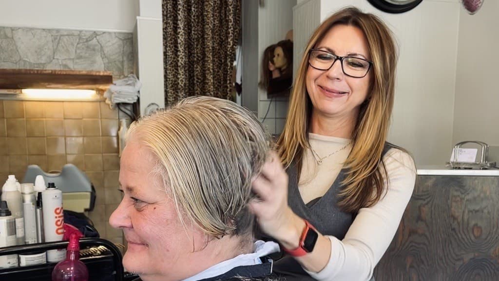 Beata Kovacs arbetar med att klippa en kunds hår i en frisörsalong. På bordet syns hårvårdsprodukter och en hårfön. Bakgrunden visar salongens inredning med kaklade väggar och gardiner.