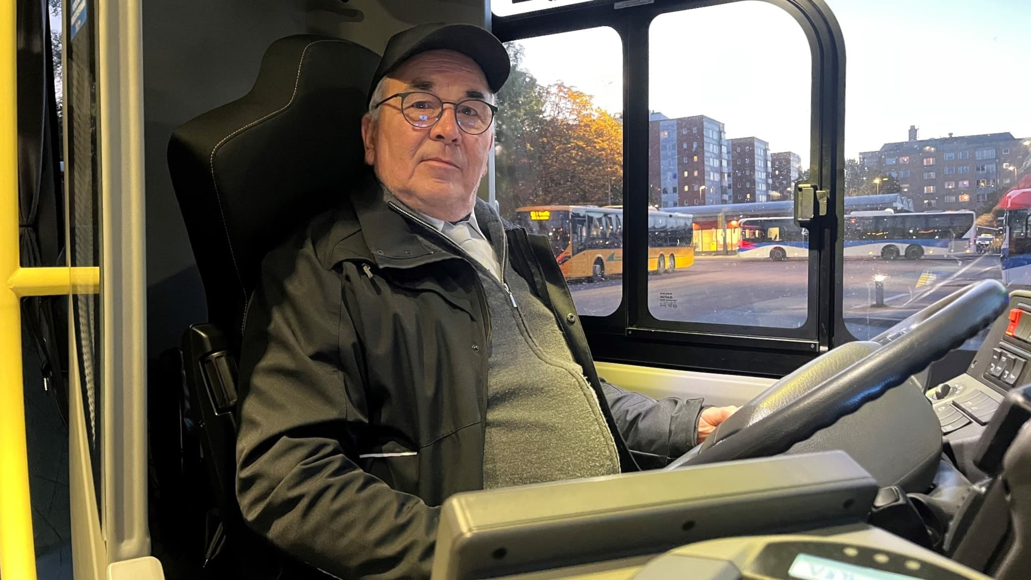 Busschauffören Olle Samuelsson sitter vid ratten i en buss, parkerad på en bussterminal. I bakgrunden syns fler bussar och byggnader i stadsmiljö under en klar himmel.