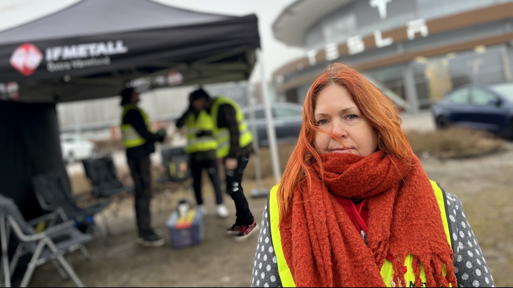 En kvinna står framför elbilstillverkaren Teslas bilhall i Örebro. Hon är klädd i en gul varselväst och en stor orange halsduk.
