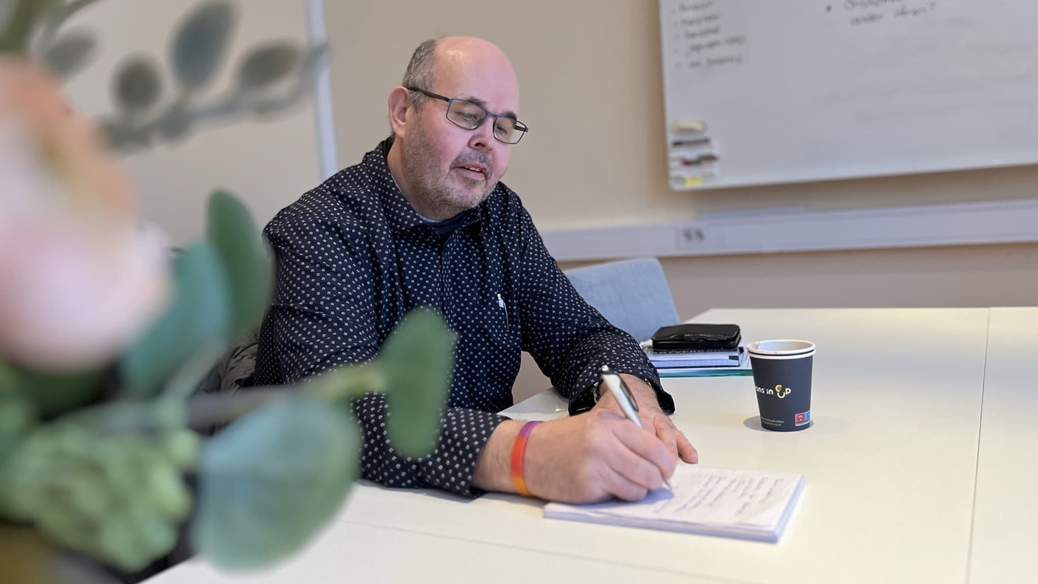 Hans-Olov Backlund, en man med delvis kalt huvud, skäggstubb, glasögon och en mörk skjorta, sitter vid ett skrivbord i ett konferensrum och skriver på ett papper. I förgrunden syns en suddig växt.