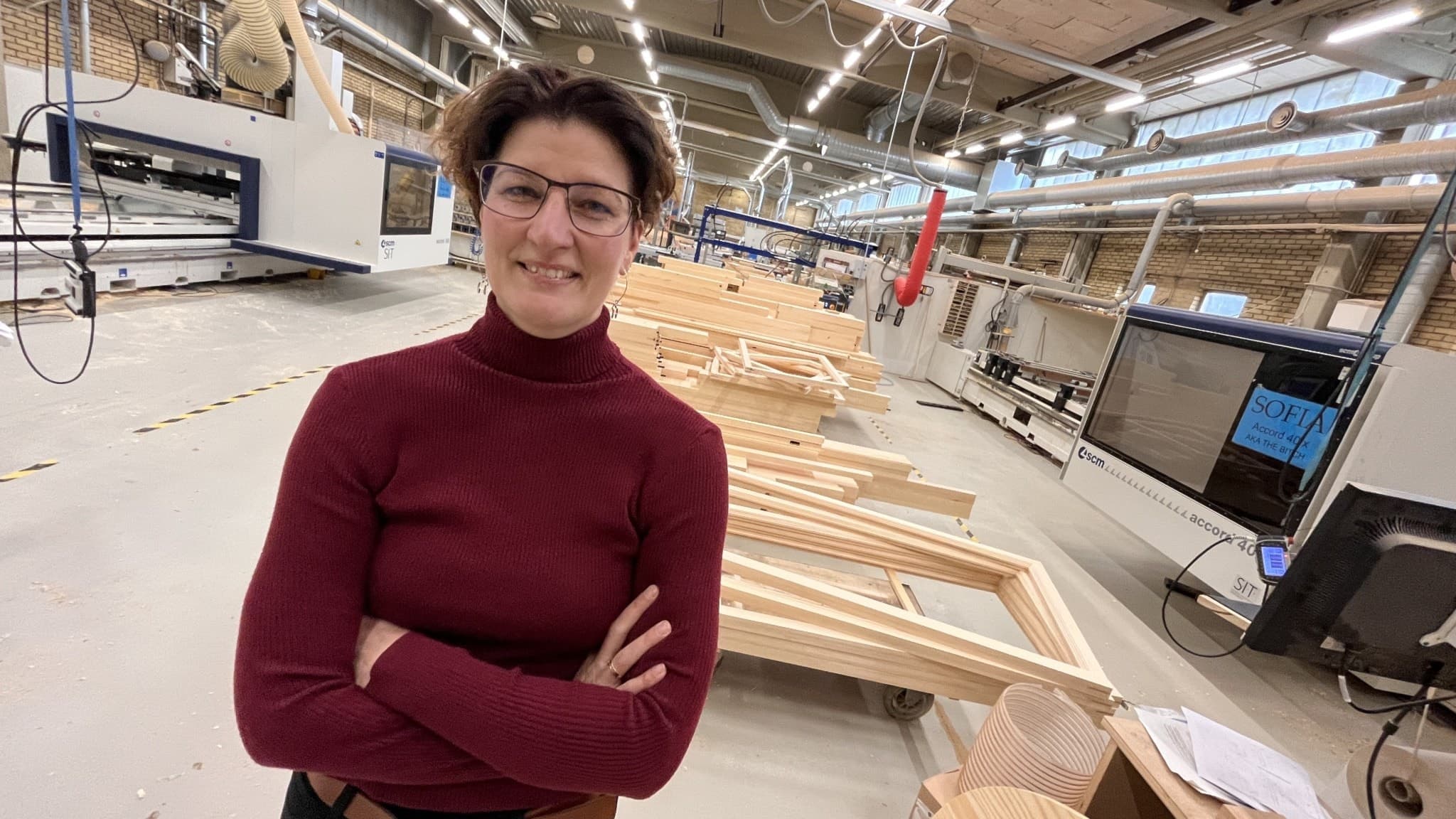 Malin Hedlund står i en modern fabriksmiljö med träprodukter och maskiner i bakgrunden, iklädd en vinröd tröja och med armarna i kors.