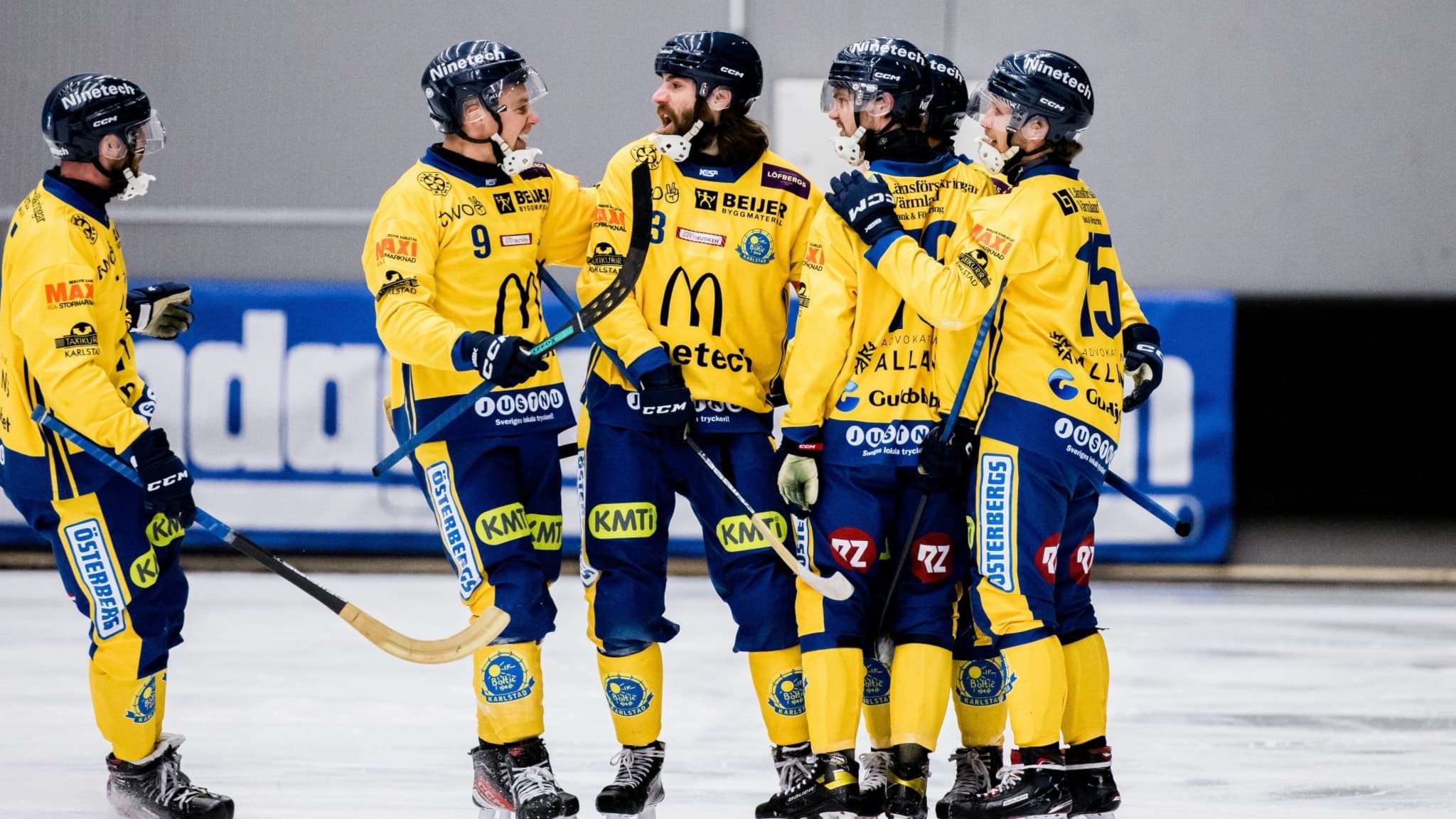 Fem bandyspelare i gula matchtröjor och blå byxor står samlade på isen under en match, med klubbor i händerna. Bakgrunden visar en reklamskylt och en grå vägg.
