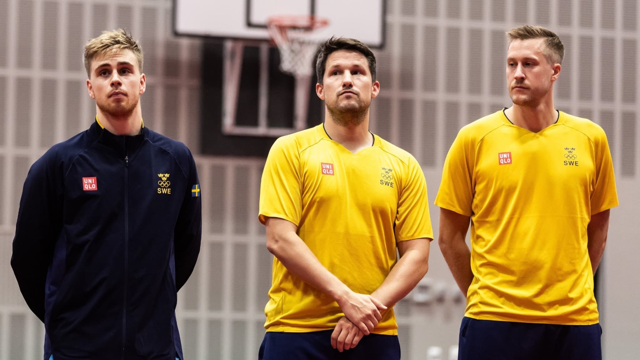 Tre män i svenska landslagets träningskläder står i en idrottshall med en basketkorg i bakgrunden. De två till höger har gula tröjor, medan mannen till vänster har en blå jacka.
