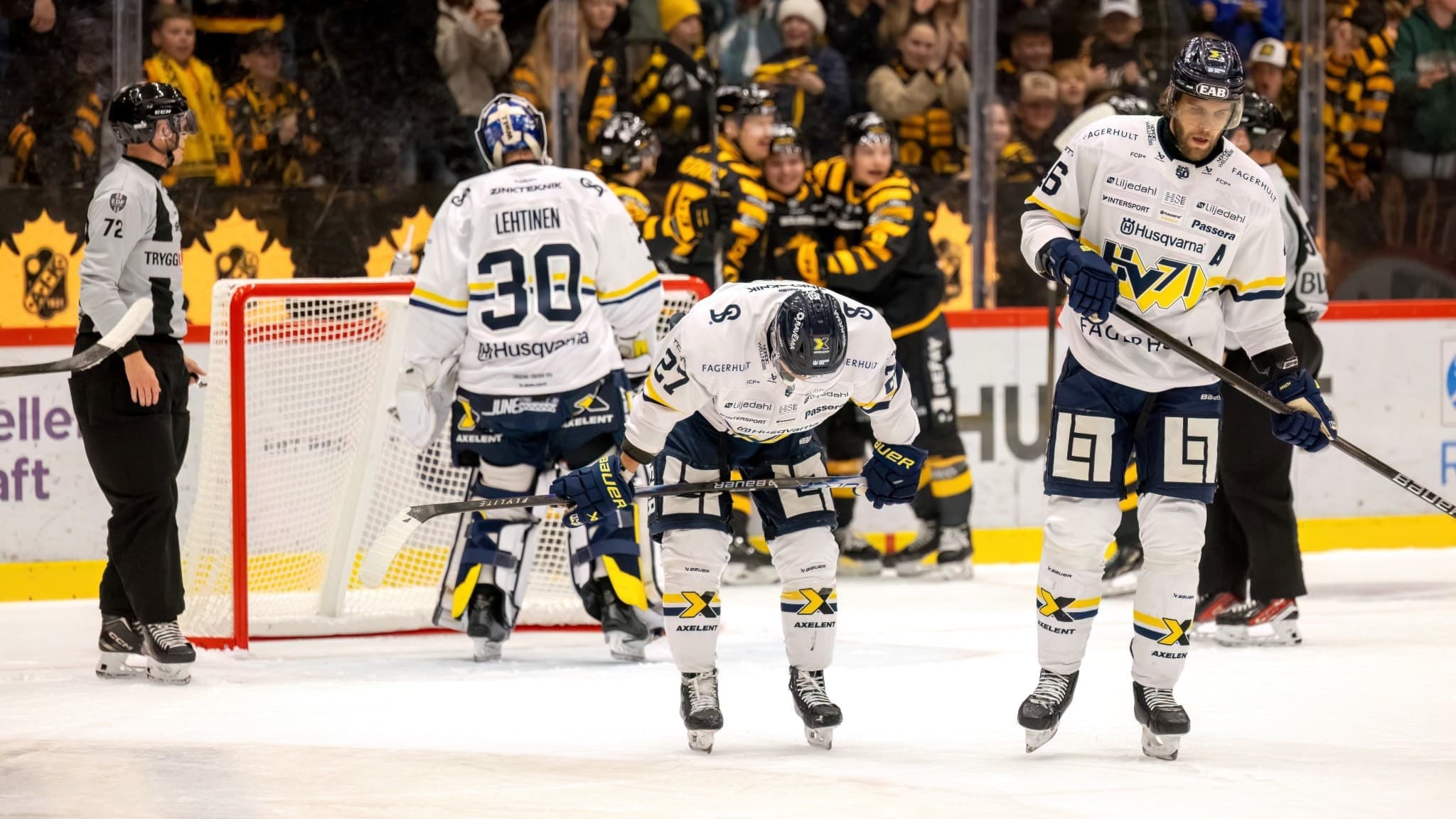Ishockeyspelare från två lag under en intensiv match, med fokus på spelare i vita tröjor framför motståndarnas målbur och publik i bakgrunden. Spänning och action på en fullsatt arena.