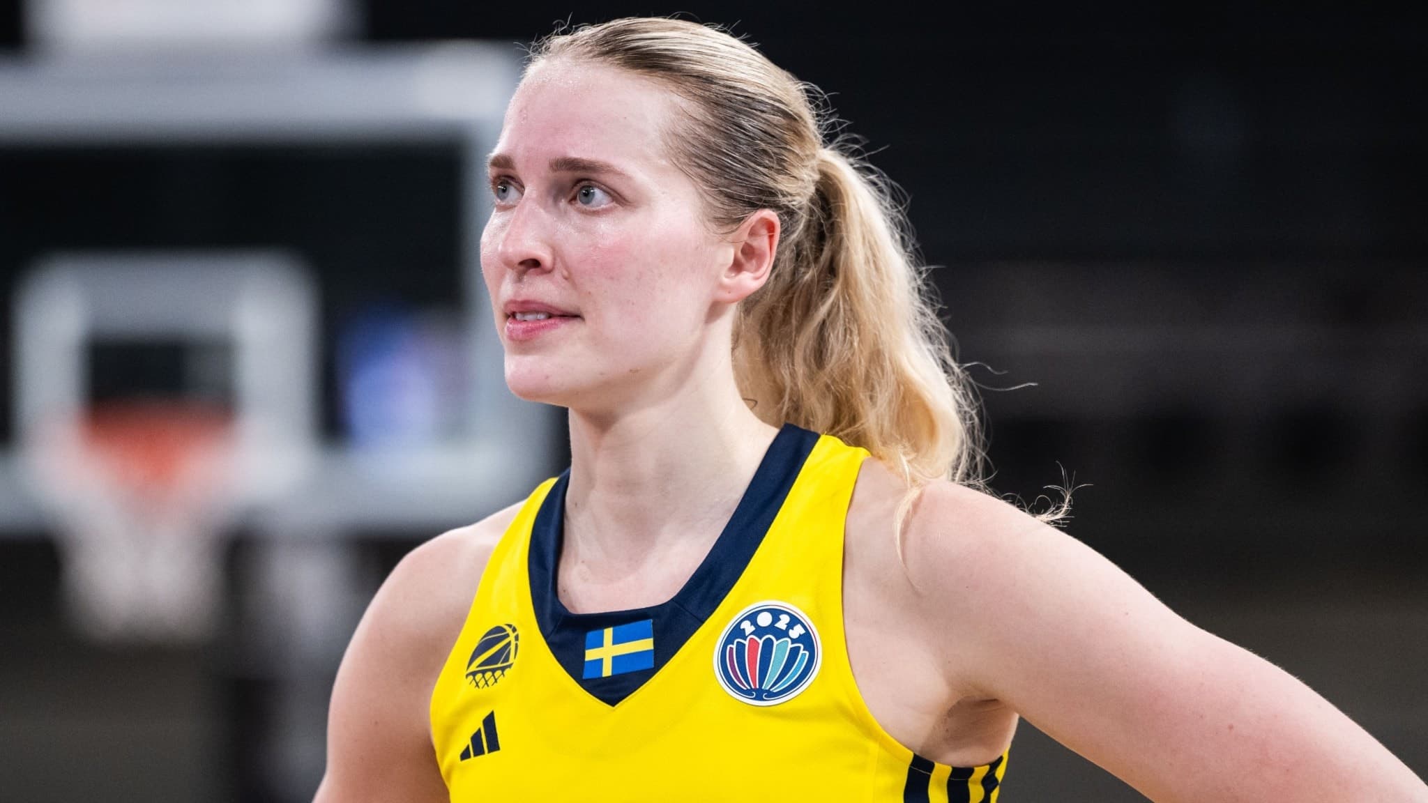 En kvinnlig basketspelare i Sveriges landslagsdräkt står på en basketplan. Hon bär en gul tröja med texten 'Sweden' och en svensk flagga syns på bröstet. Basketkorg i bakgrunden.