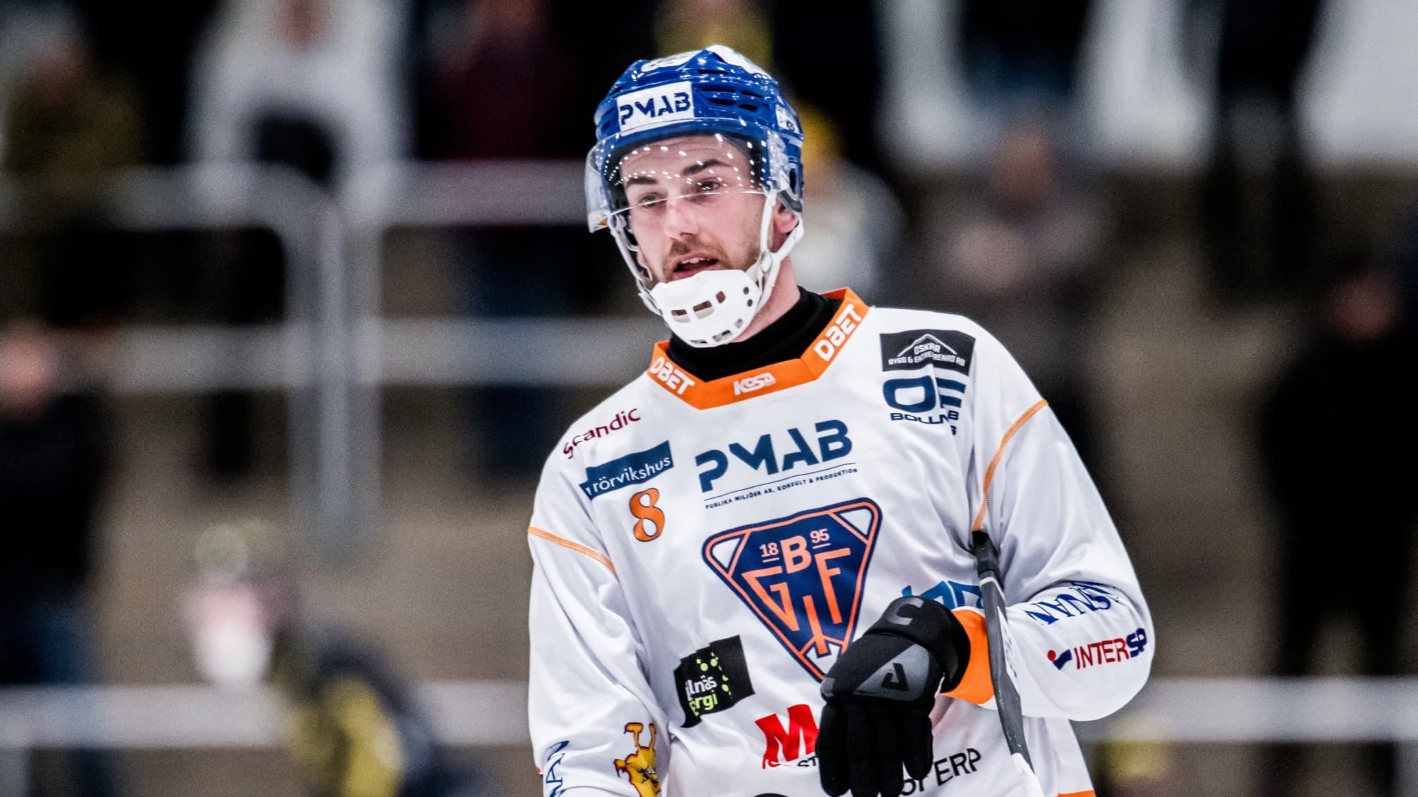 En ishockeyspelare i vita och orangea matchkläder med PMAB och HV71-logotyp syns på isen under en match. Bakgrunden visar åskådare i en ishall.