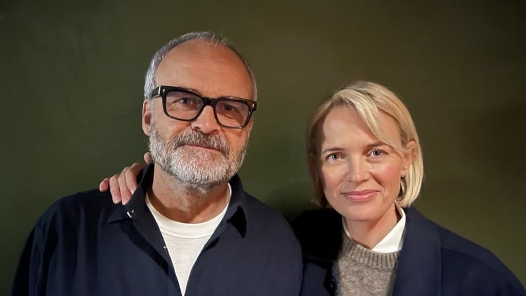 Två personer står bredvid varandra framför en mörkgrön bakgrund, båda klädda i mörka skjortor och tröjor. Den ena personen har grått hår, medan den andra har blont hår.