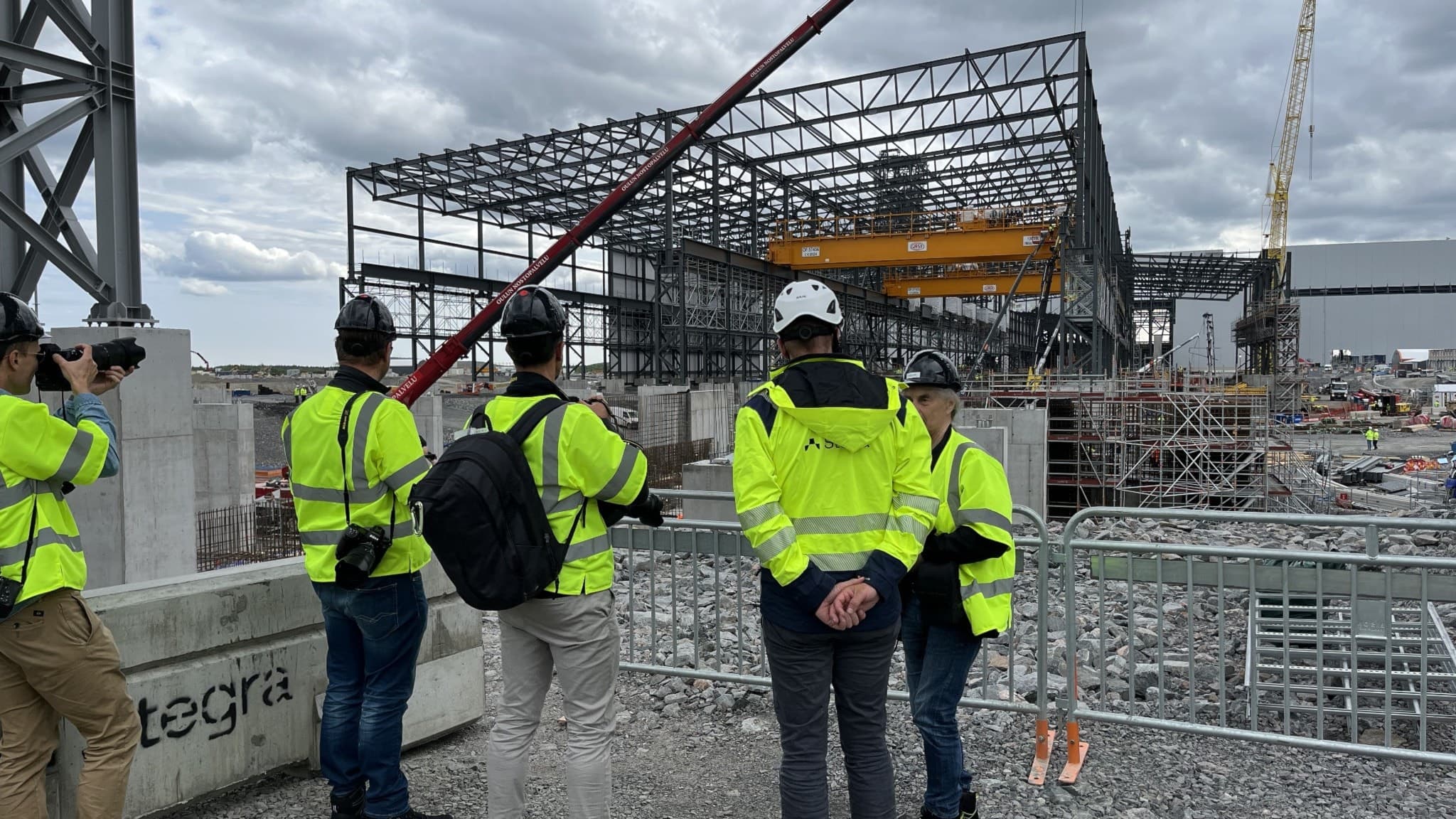 Fyra personer i gula reflexvästar och hjälmar står vid en byggarbetsplats och tittar på en stålstruktur under uppbyggnad. Mörka moln syns i bakgrunden, vilket skapar en dramatisk atmosfär.
