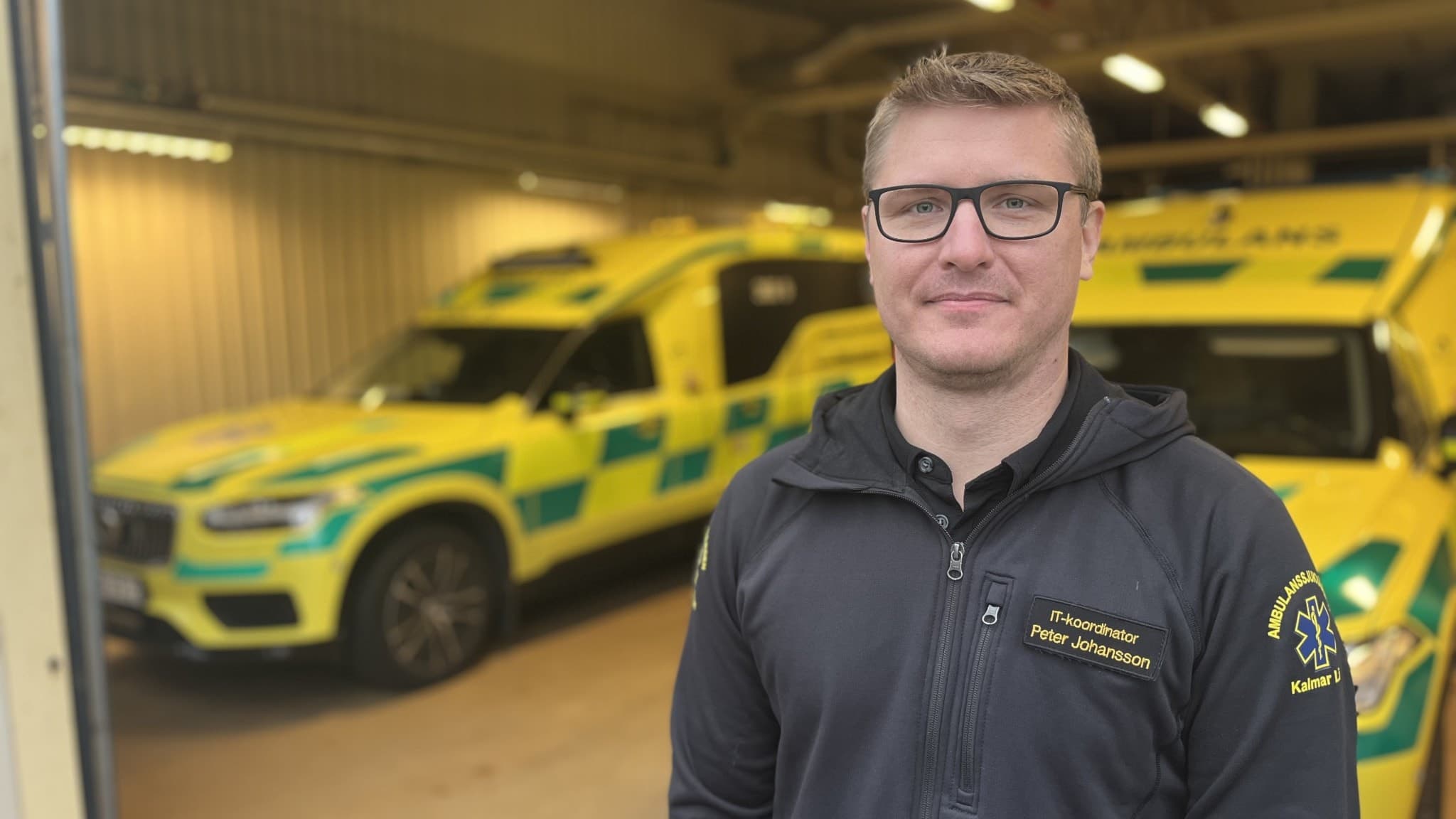 Peter Johansson, it-koordinator för ambulanssjukvården i Region Kalmar län, står framför två gula ambulansfordon i en garagebyggnad, klädd i en mörk hoodie.