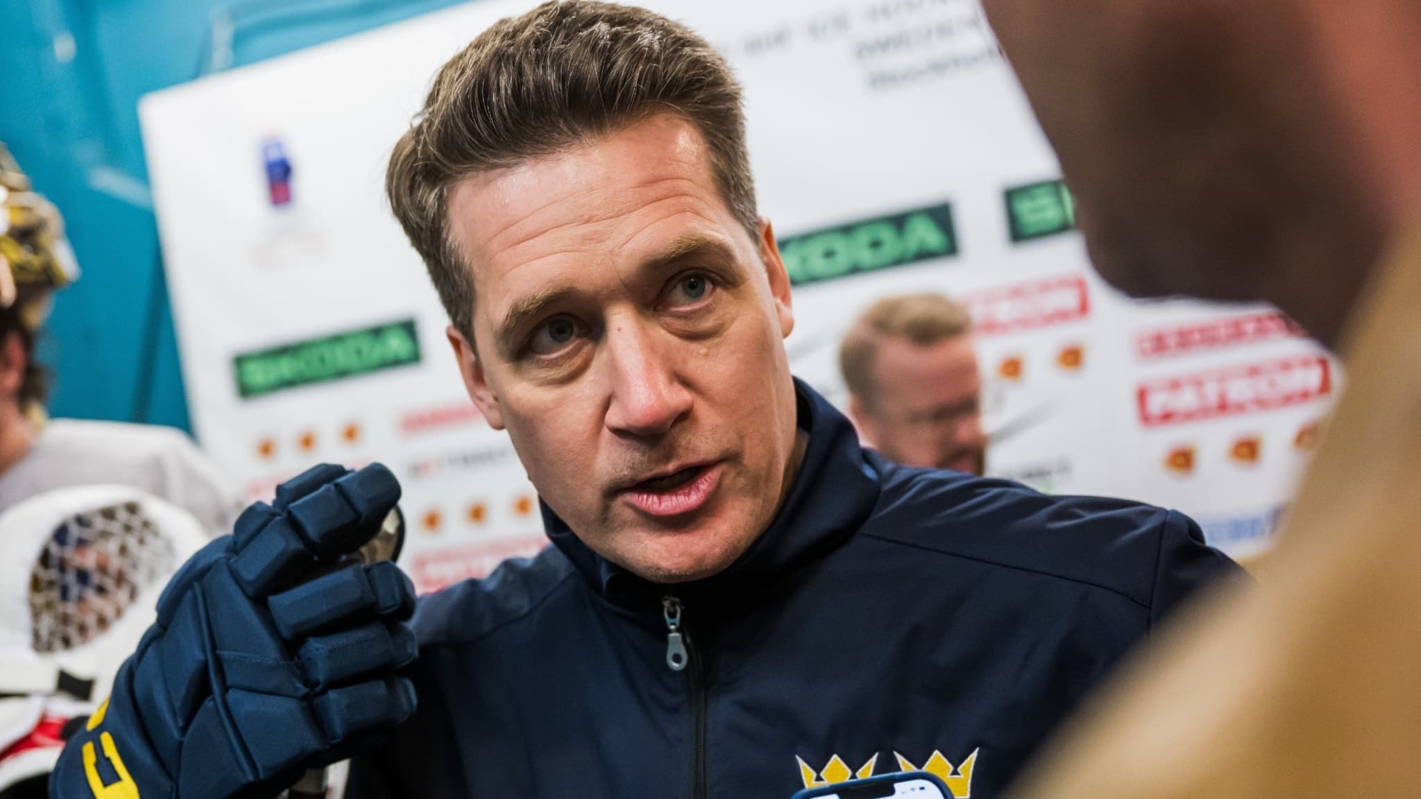 En person i mörk sportjacka med Sverigelogo på bröstet håller en mikrofon framför sig. I bakgrunden syns en intervjuvägg med logotyper från sponsorer som Skoda och Svenska Spel.
