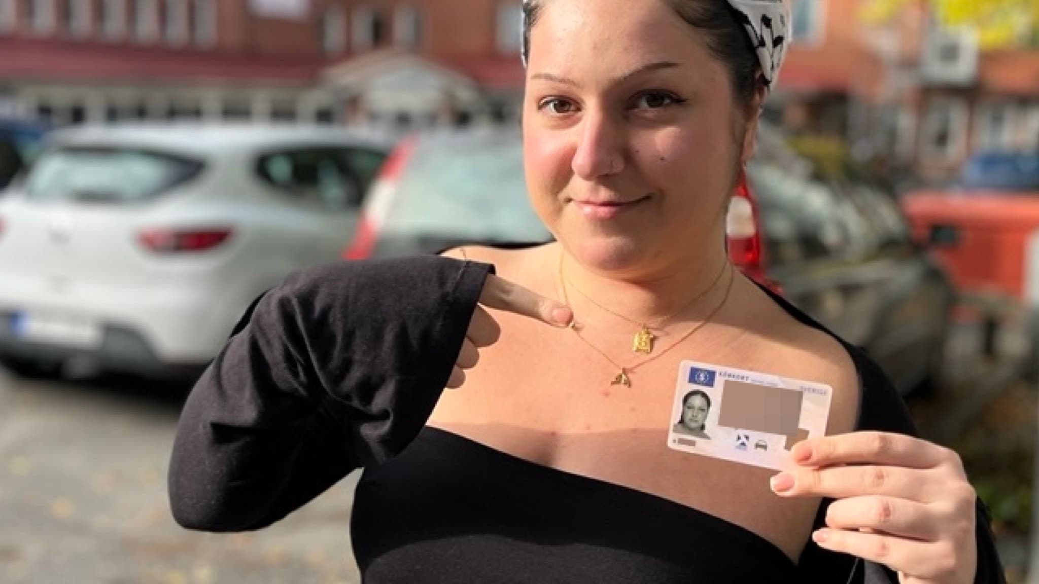 En person håller upp ett ID-kort utomhus på en parkeringsplats med flera bilar och en tegelbyggnad i bakgrunden. Personen är klädd i en svart tröja och har ett mönstrat hårband.