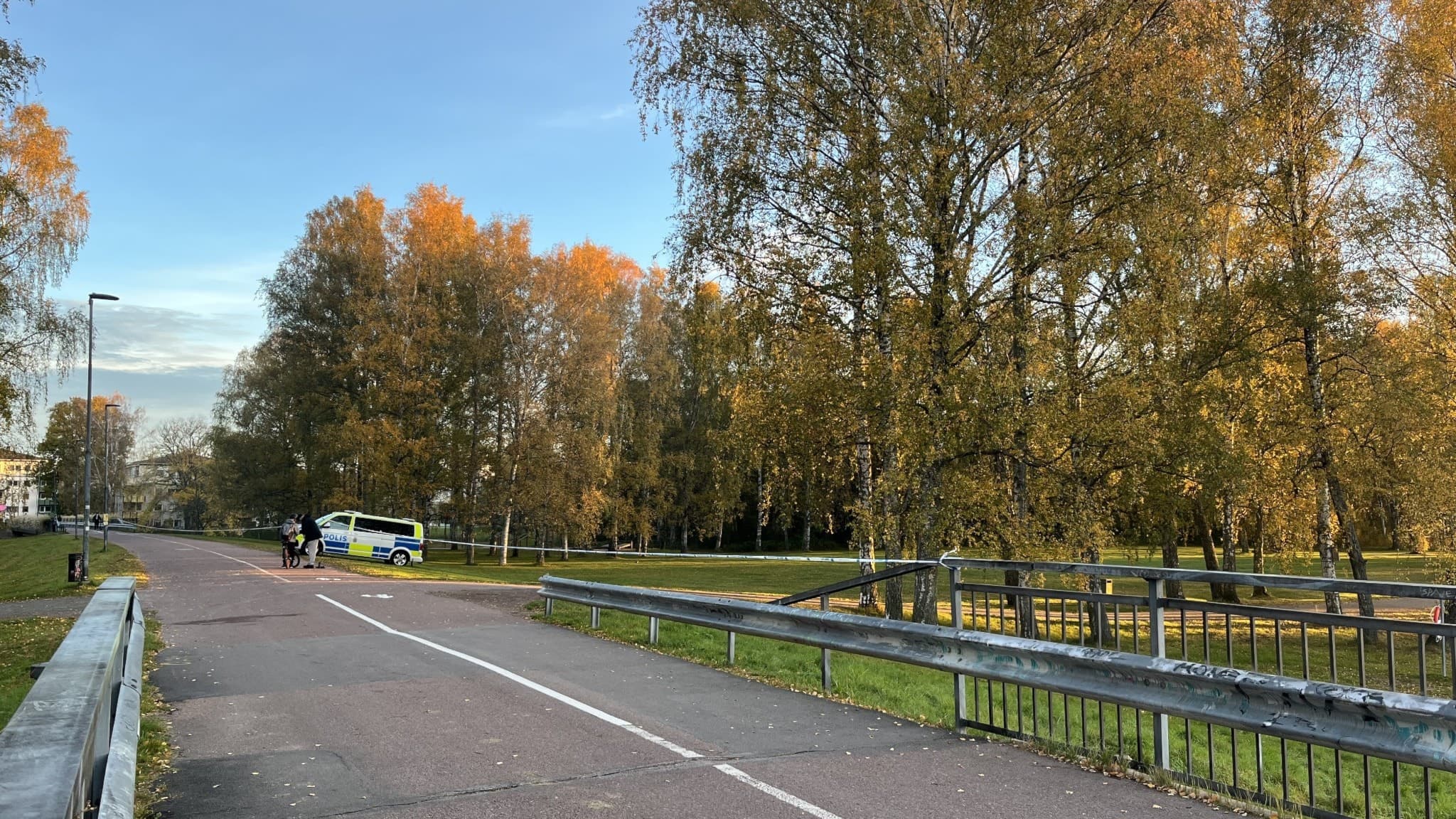 Avspärrad gångväg vid Gubbholmen i Karlstad, med polisnärvaro. Höstfärgade träd längs vägen och en polisbil synlig i bakgrunden. Händelsen verkar pågå i ett lugnt område.