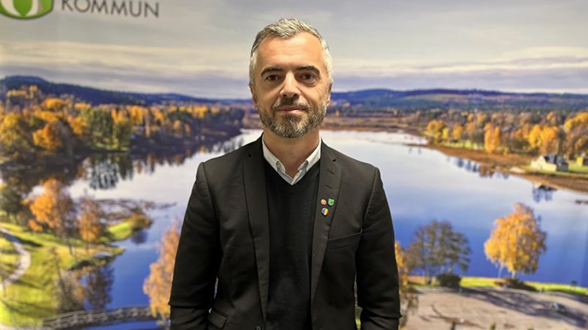 Politikern Driton Bilalli (S) står i kostym framför en bild på Tranemo.
