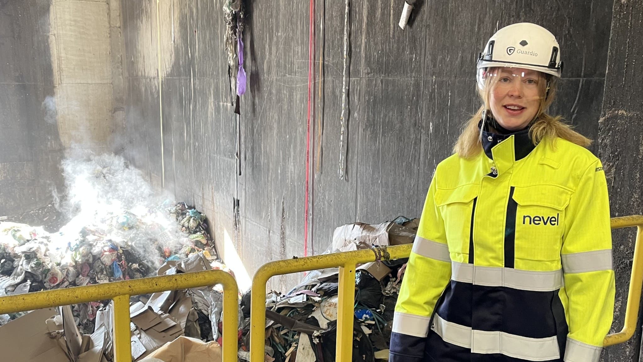 En kvinna i varselkläder och hjälm står framför ett staket med stora mängder avfall i bakgrunden.