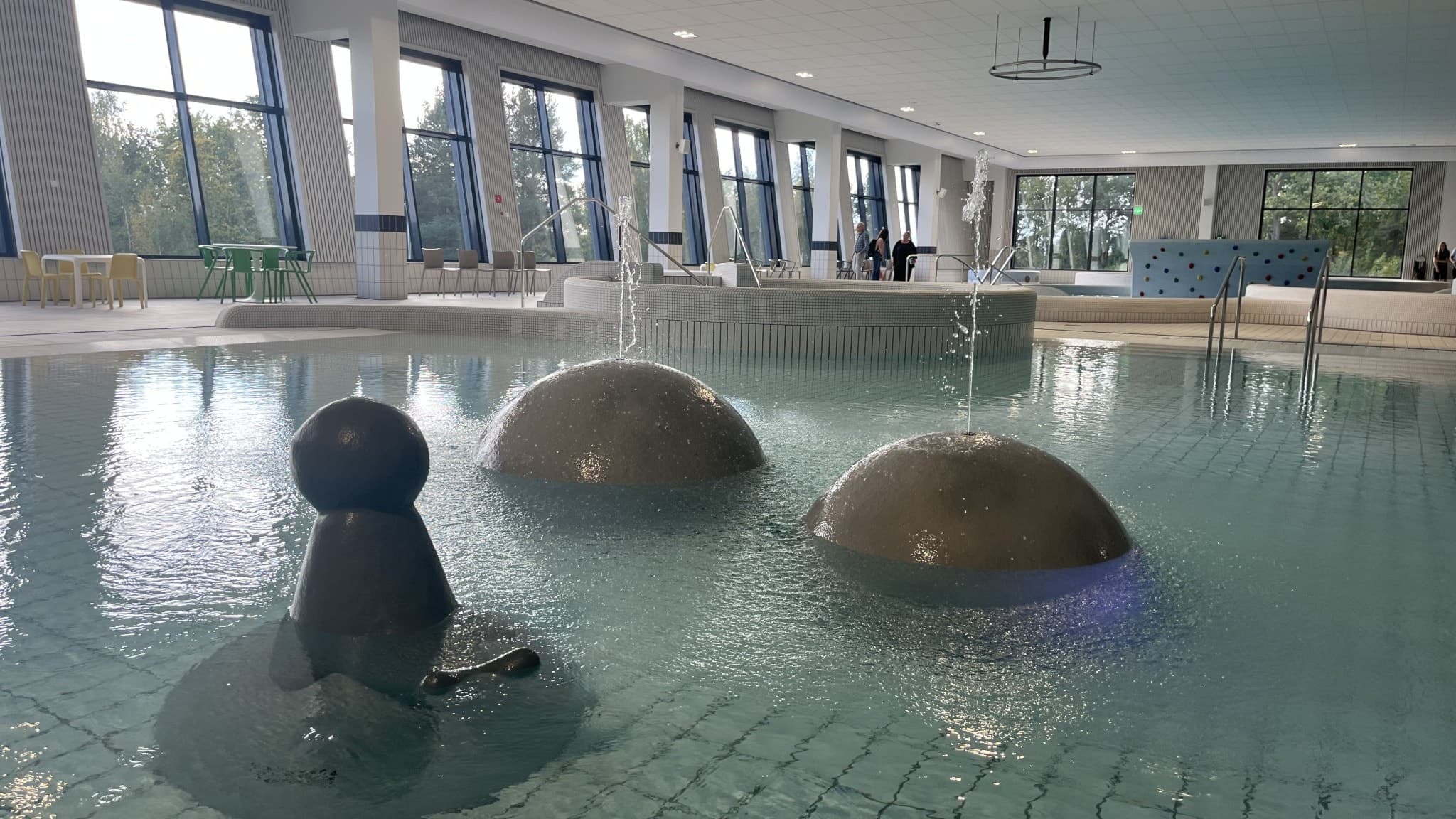 Plask- och badbassäng i simhall inomhus. Runda skulpturer med vattenstrålar mitt i bassängen. 