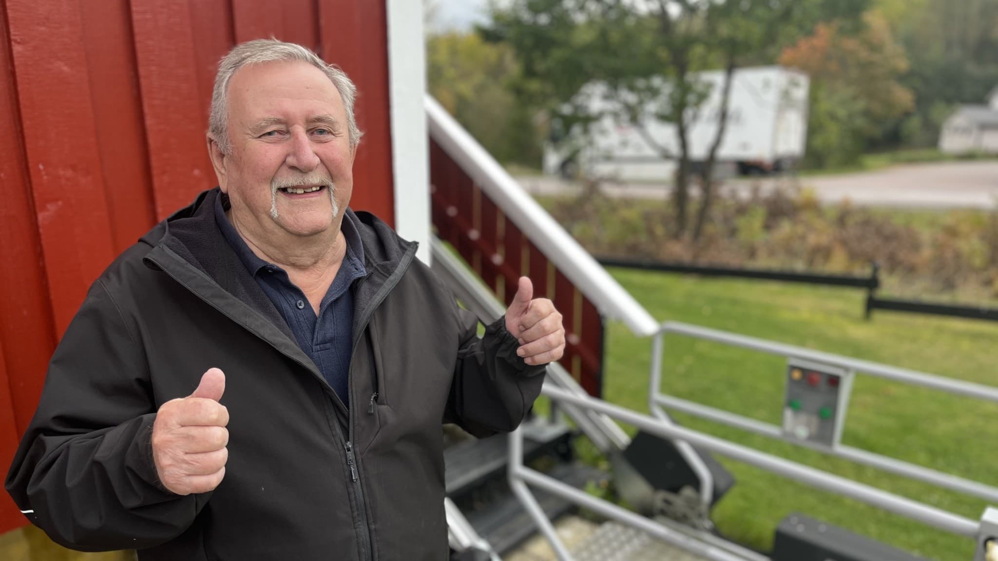 Tore Olsson står framför sitt röda hus nära E45 med tummen upp, glad över Trafikverkets nya plan som låter honom bo kvar. Bakgrunden visar en grönskande miljö och en parkerad bil.