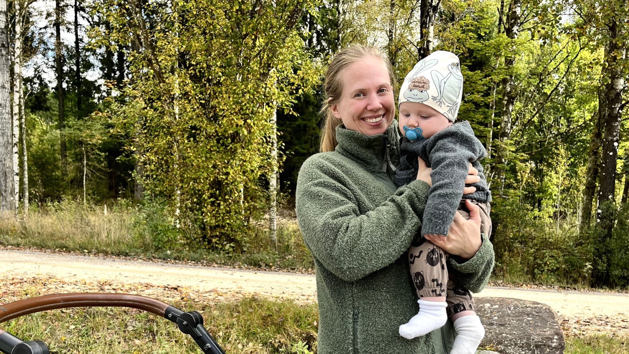 Tvåbarnsmamman Ida Hermansson står utomhus med sitt barn, Folke 5 månader, på armen, omgiven av grönska. Framför henne syns en barnvagn på en grusväg i en naturskön miljö med träd i bakgrunden.