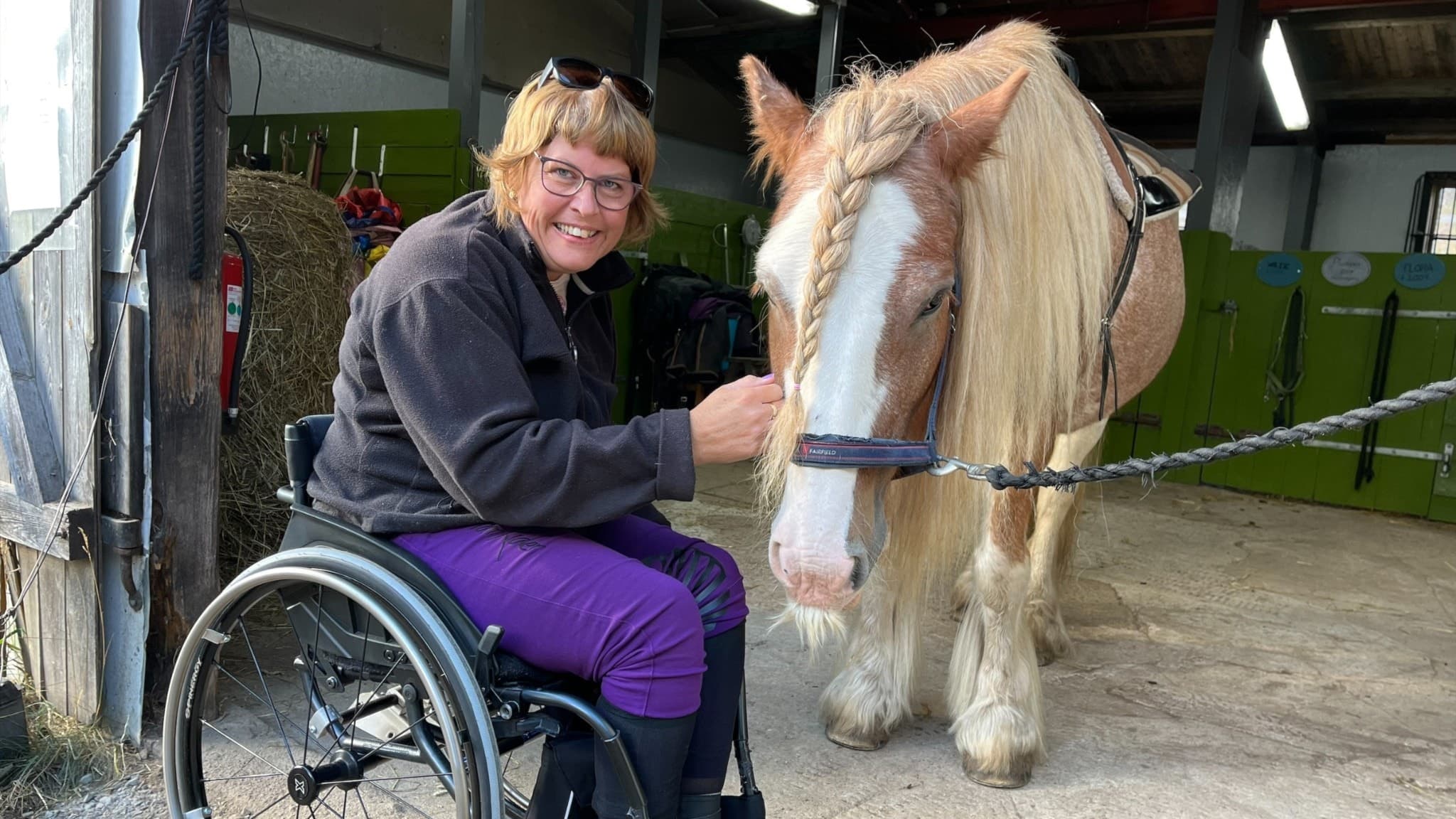 Petra Lennartsson i rullstol klappar en stor häst med lång man i ett stall. Hästen står fastbunden, och miljön präglas av stallinredning och halvdunkel belysning.