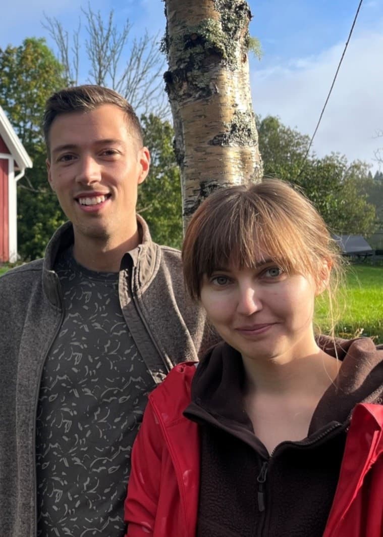 Elin och Sebastian står framför sitt nyinköpta röda hus med vita knutar, omgivna av grönska. I bakgrunden syns en grusväg, staket och ett träd i soligt väder.
