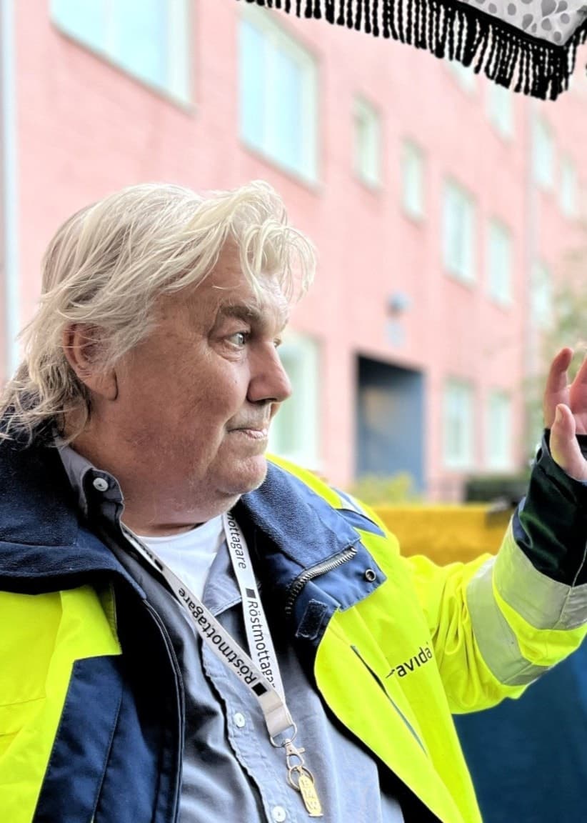 Mikael Sandbacka, klädd i gul varseljacka, gestikulerar med handen under ett tak utomhus. Bakgrunden visar en byggnad med röda tegelväggar och grönskande omgivning.