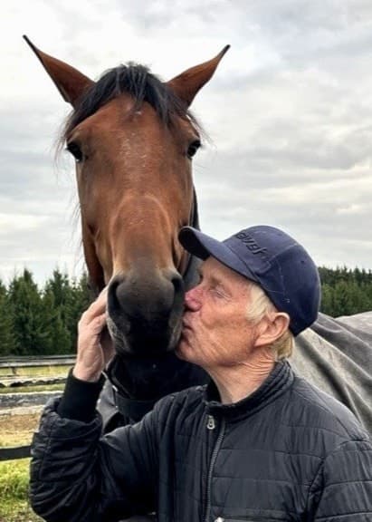 Stefan Melander står bredvid en brun häst med täcke på en hästgård, omgiven av staket och en skog i bakgrunden under en molnig himmel.