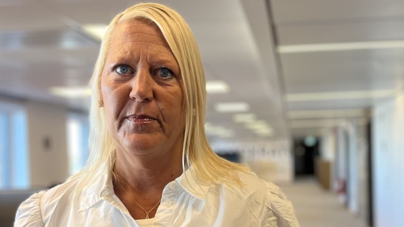 En kvinna i vit blus står inomhus med suddad bakgrund. Till höger syns en hand som håller ett valkuvert framför en blå bakgrund med röd detalj.