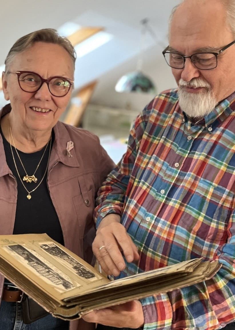 Gunilla och Benny Kullinger står i ett ljust rum och bläddrar i ett nyinköpt vykortsalbum. I bakgrunden syns möbler och utställningsföremål.