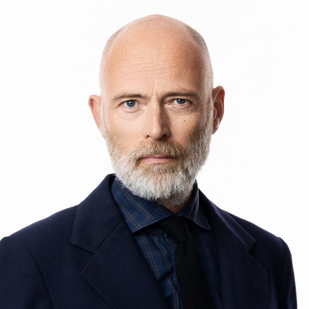 Knut Kainz Rognerud, klädd i mörk kostym, ses i förgrunden under en presskonferens om ekonomipriset i en elegant sal med kristallkronor och guldinramade porträtt.