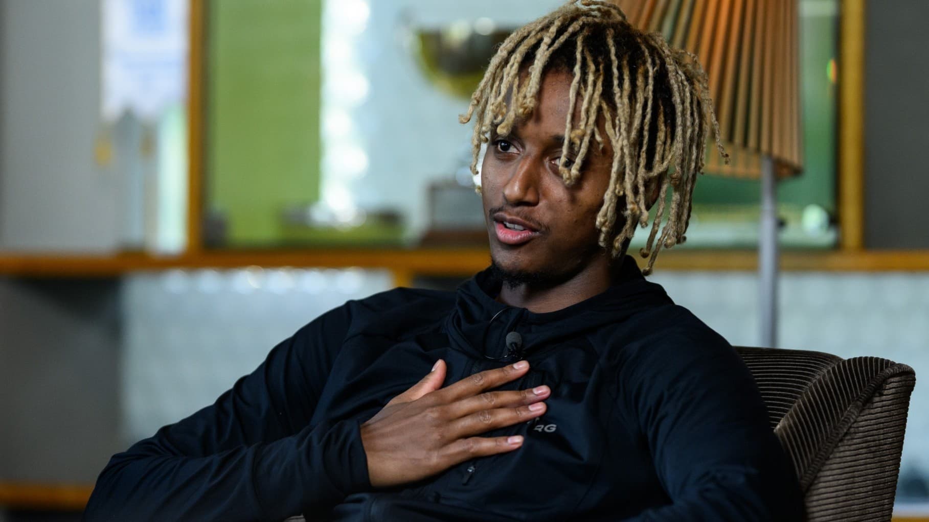 En person med ljusa dreadlocks sitter i en fåtölj med en hand på bröstet, iklädd en mörk tröja. Bakgrunden visar en modern inomhusmiljö med en lampa och glasdetaljer.