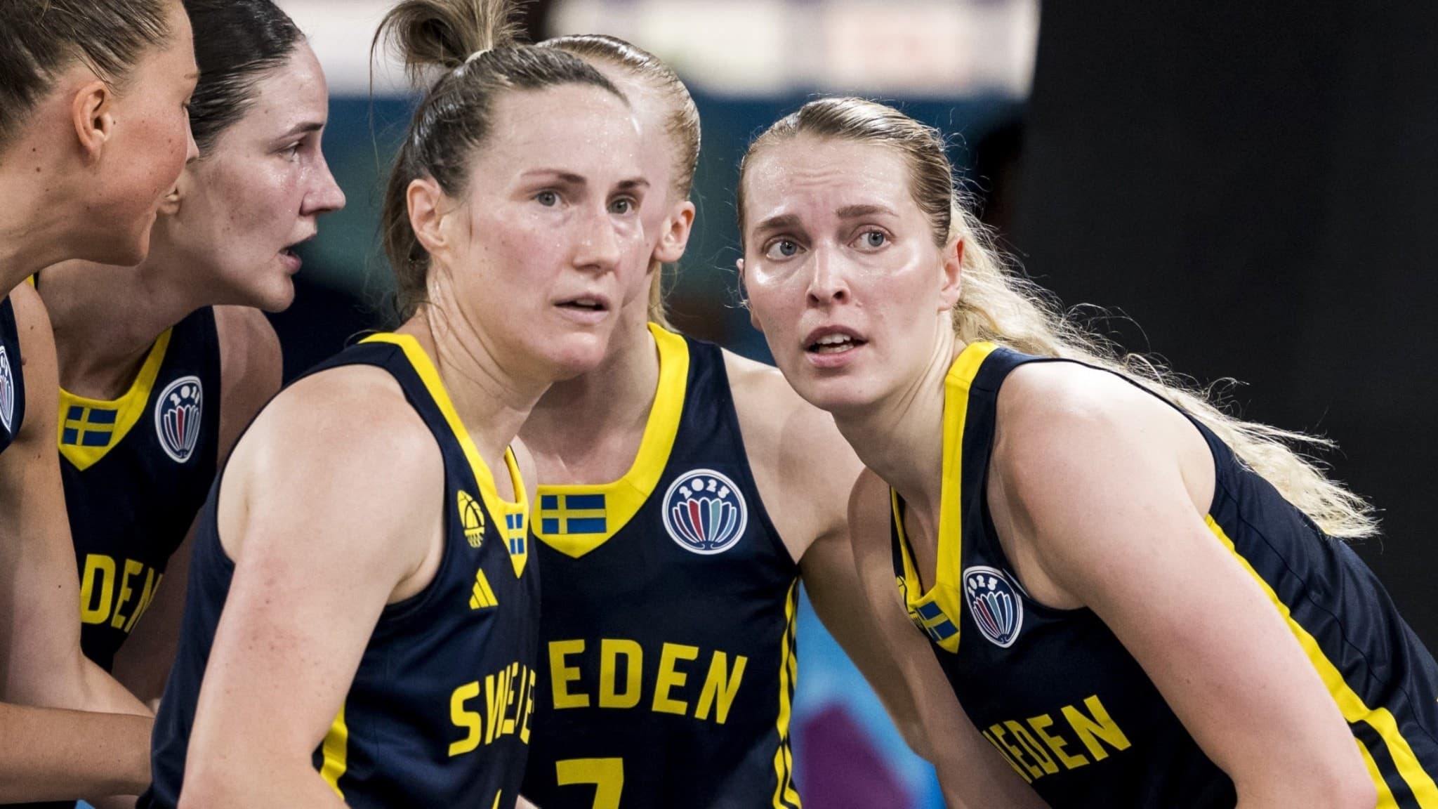 Svenska basketspelare i landslagsdräkter med texten 'Sweden' och svenska flaggan synlig, samlade under en match i en inomhusarena.